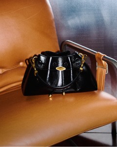 Black patent leather drawstring bag with gold-tone hardware displayed on a tan leather chair.