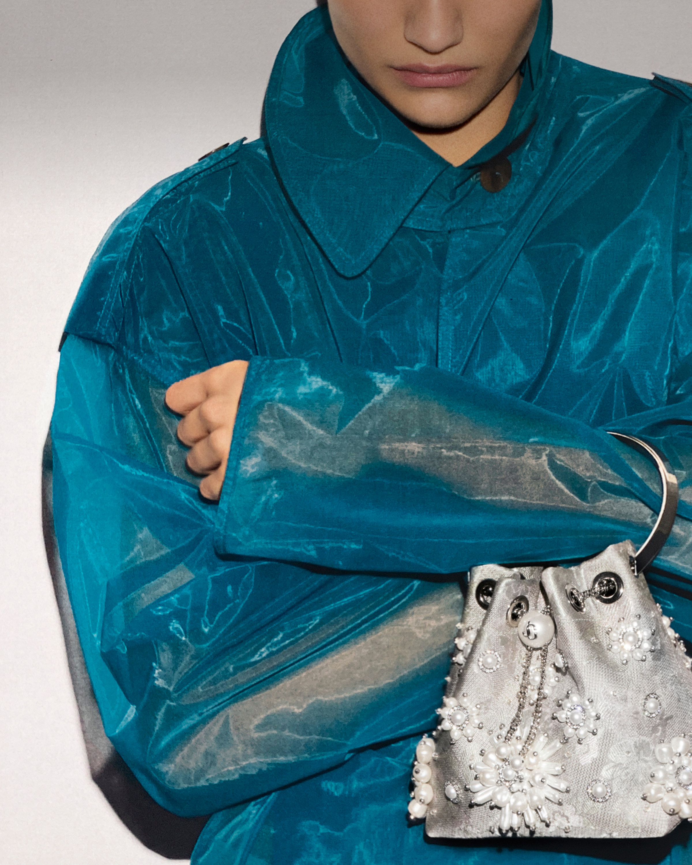 Model wearing a turquoise coat and holding a drawstring bag with pearl and crystal embellishments.