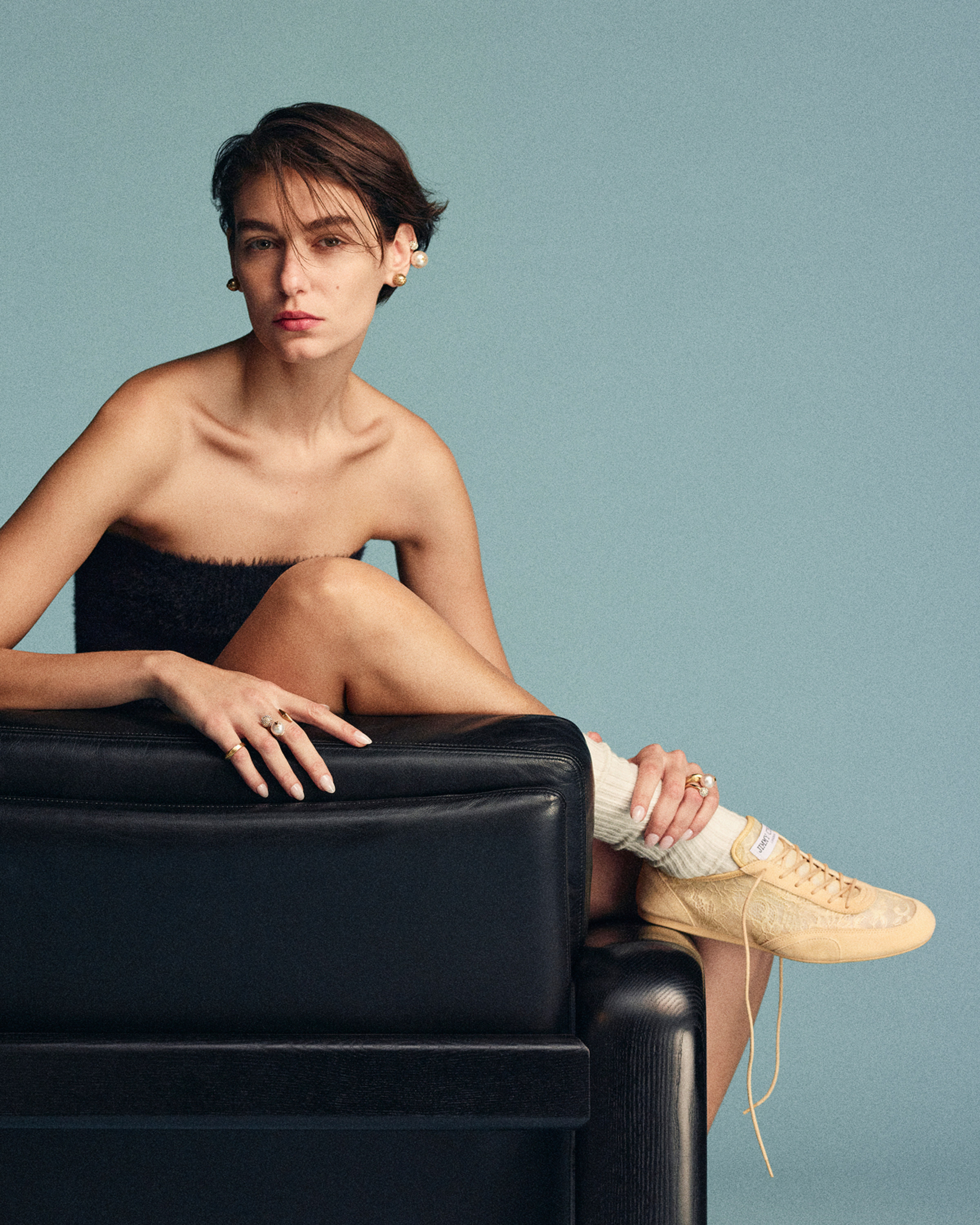 Model seated on a black chair wearing yellow lace low-top trainers against a blue background.