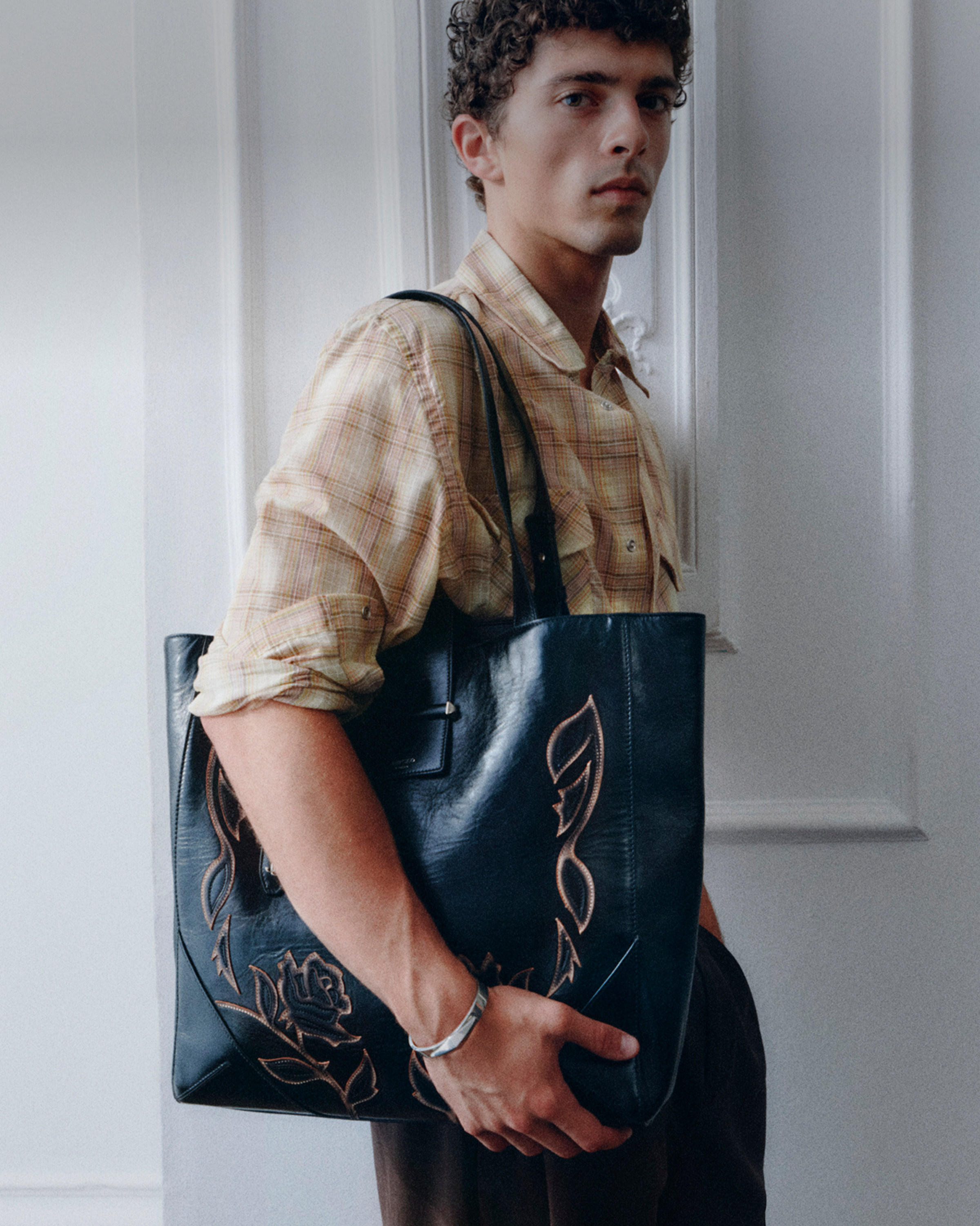 Rory Macleod wearing a patterned neutral shirt and carrying a black leather tote bag with floral detailing.