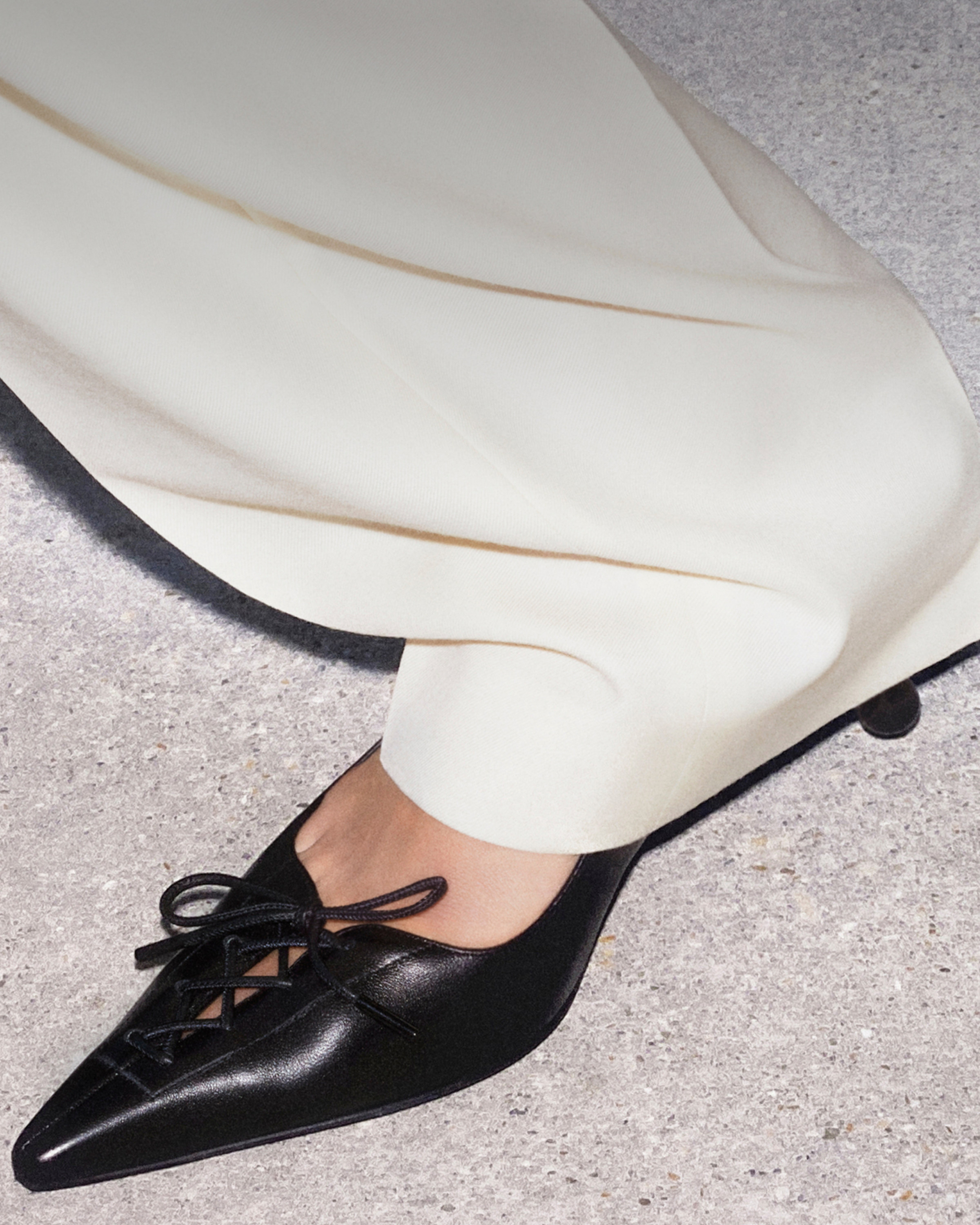 Close-up of black patent leather pointed corset lace-up shoes worn with cream trousers.