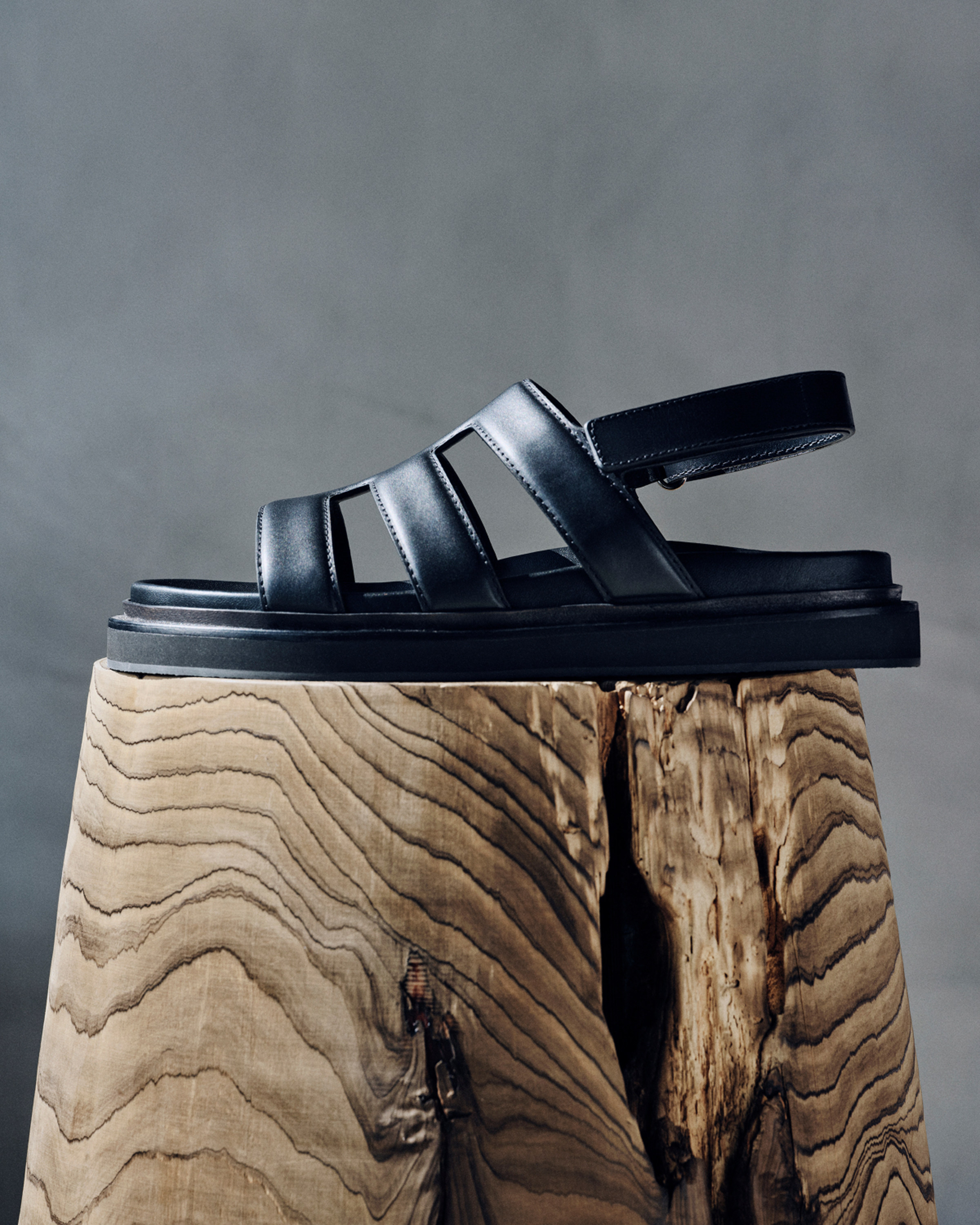Black leather strap sandal with chunky sole displayed on a wooden block.