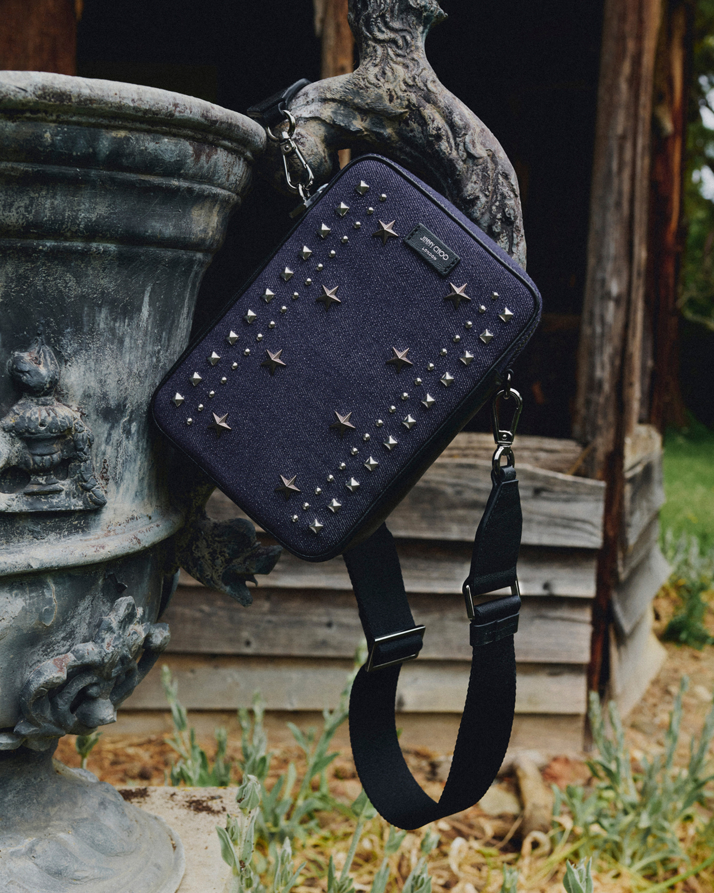Navy crossbody bag with star and stud embellishments suspended beside a decorative stone urn.