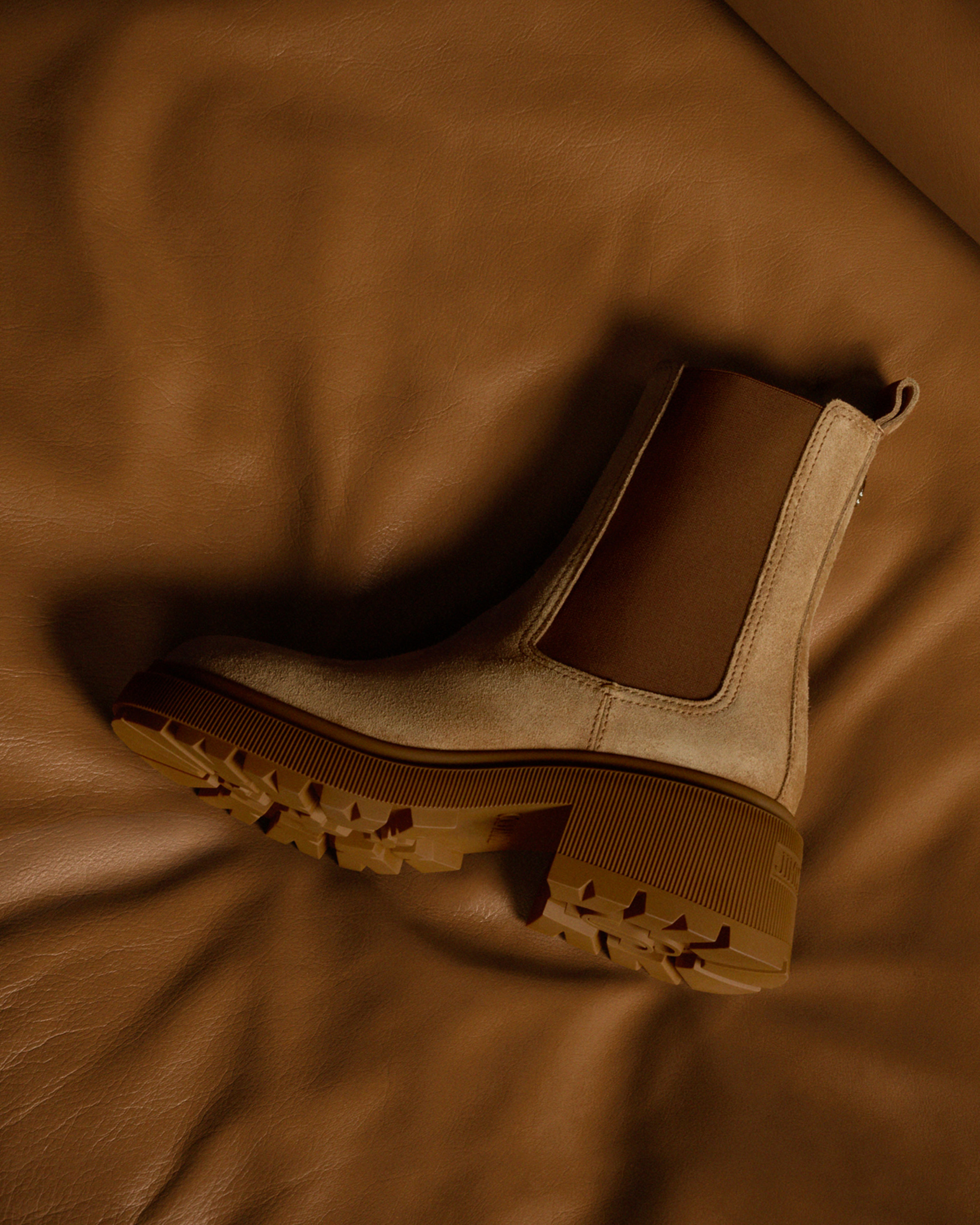 Beige suede Chelsea boot with elasticated side panels and a chunky lug sole, displayed on a brown leather surface.
