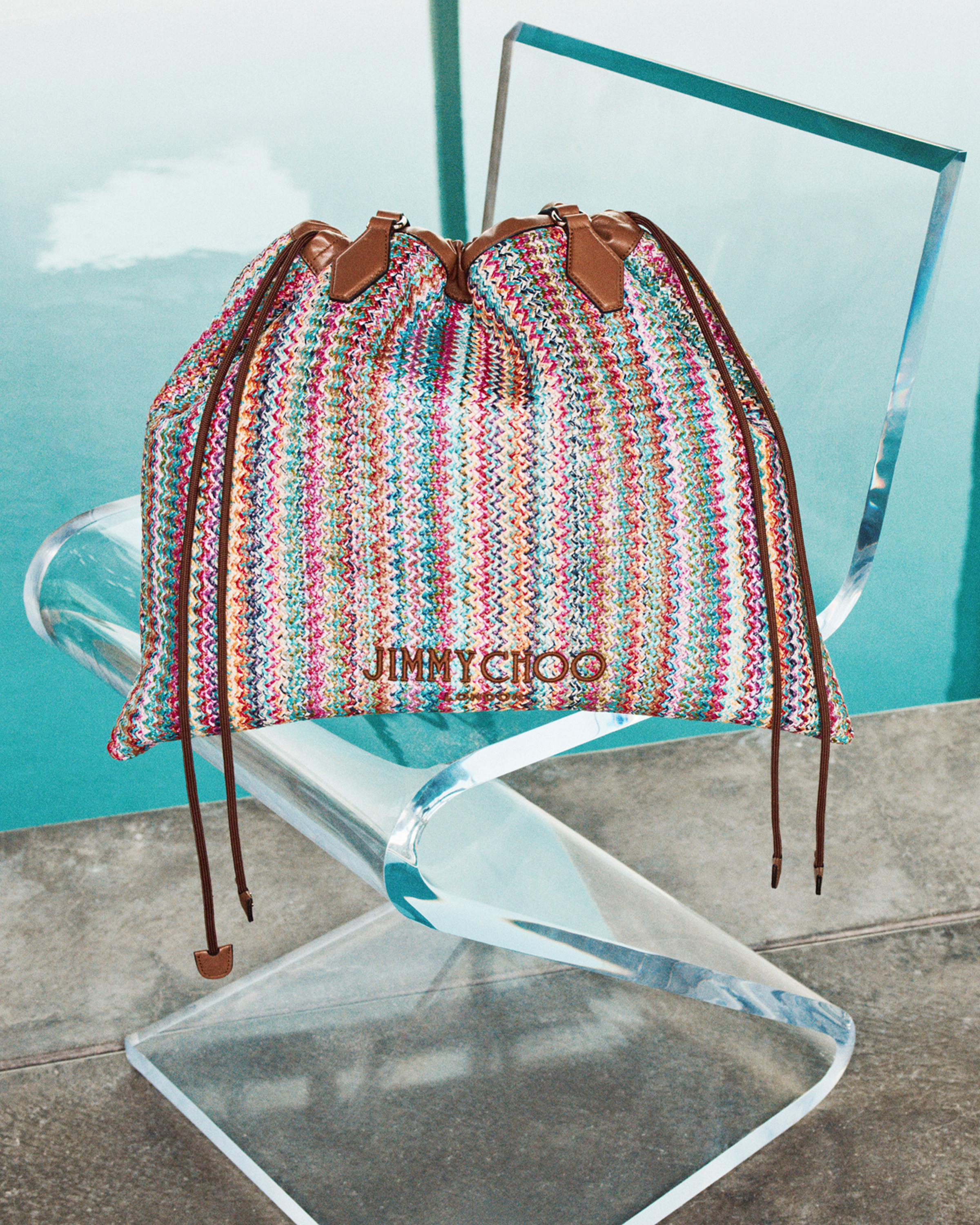 Multicolour woven drawstring bag with leather ties placed on a clear chair by a pool.