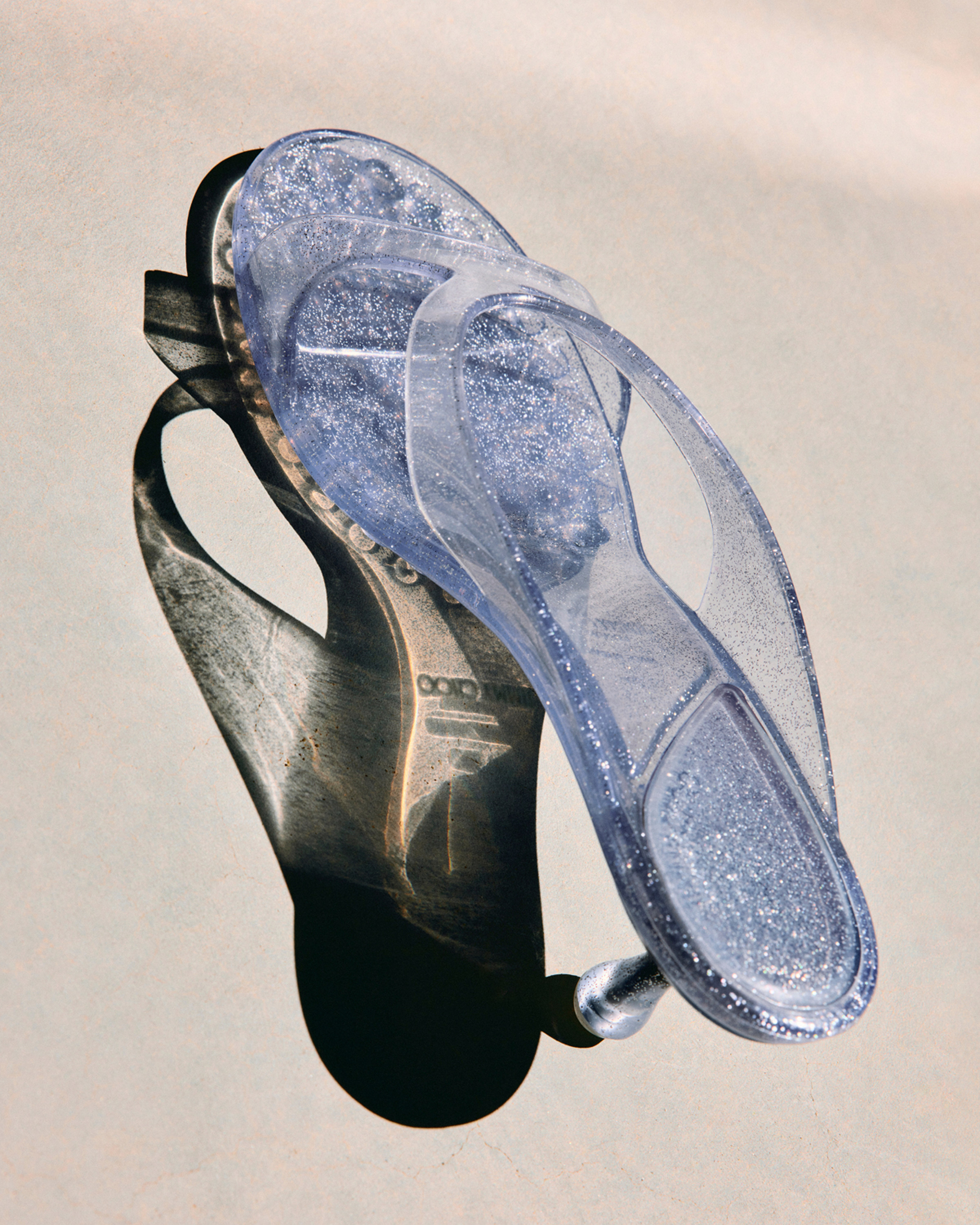 Transparent glitter jelly sandal with sculptural heel casting a shadow in sunlight.