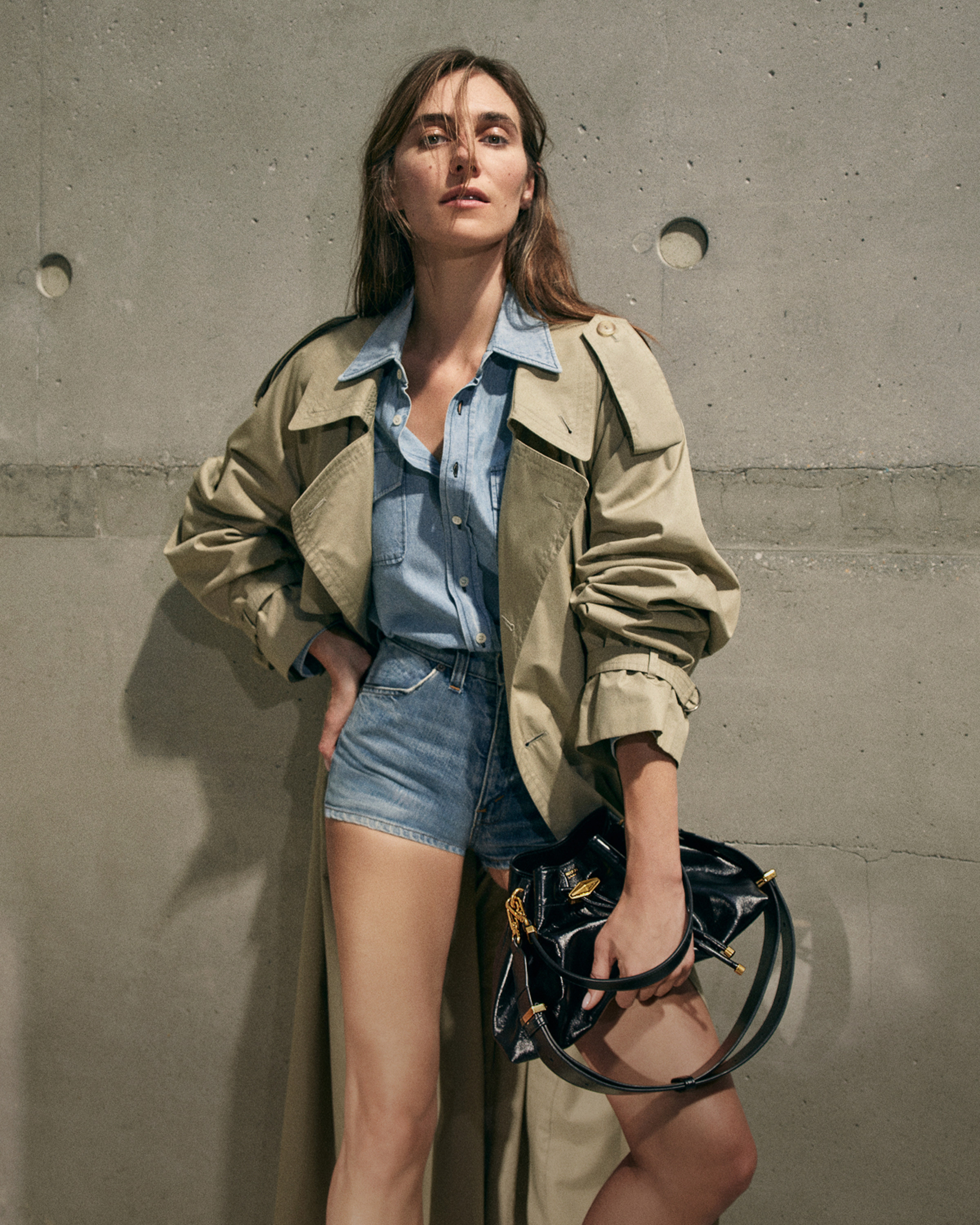 Devon Lee Carlson in a beige trench coat and denim outfit holding a black patent leather drawstring bag with gold-tone hardware.