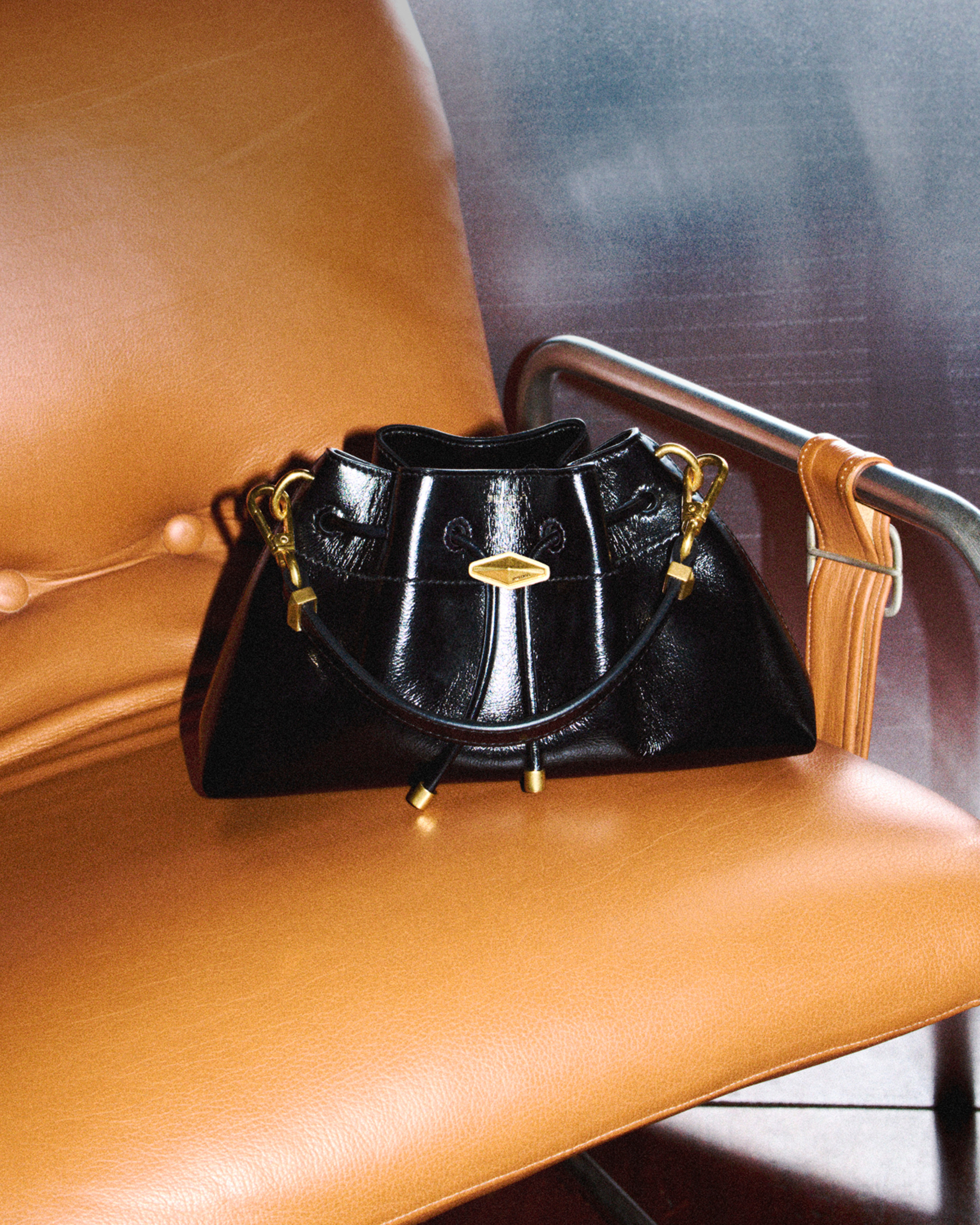 Black shiny leather drawstring bag with gold-tone hardware displayed on a tan leather chair.