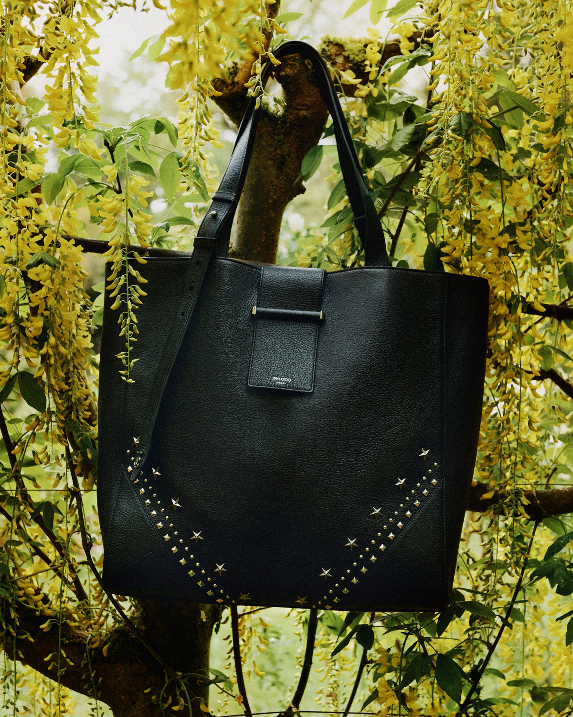 Black leather tote bag with stud detailing hanging from a tree among cascading yellow blossoms.