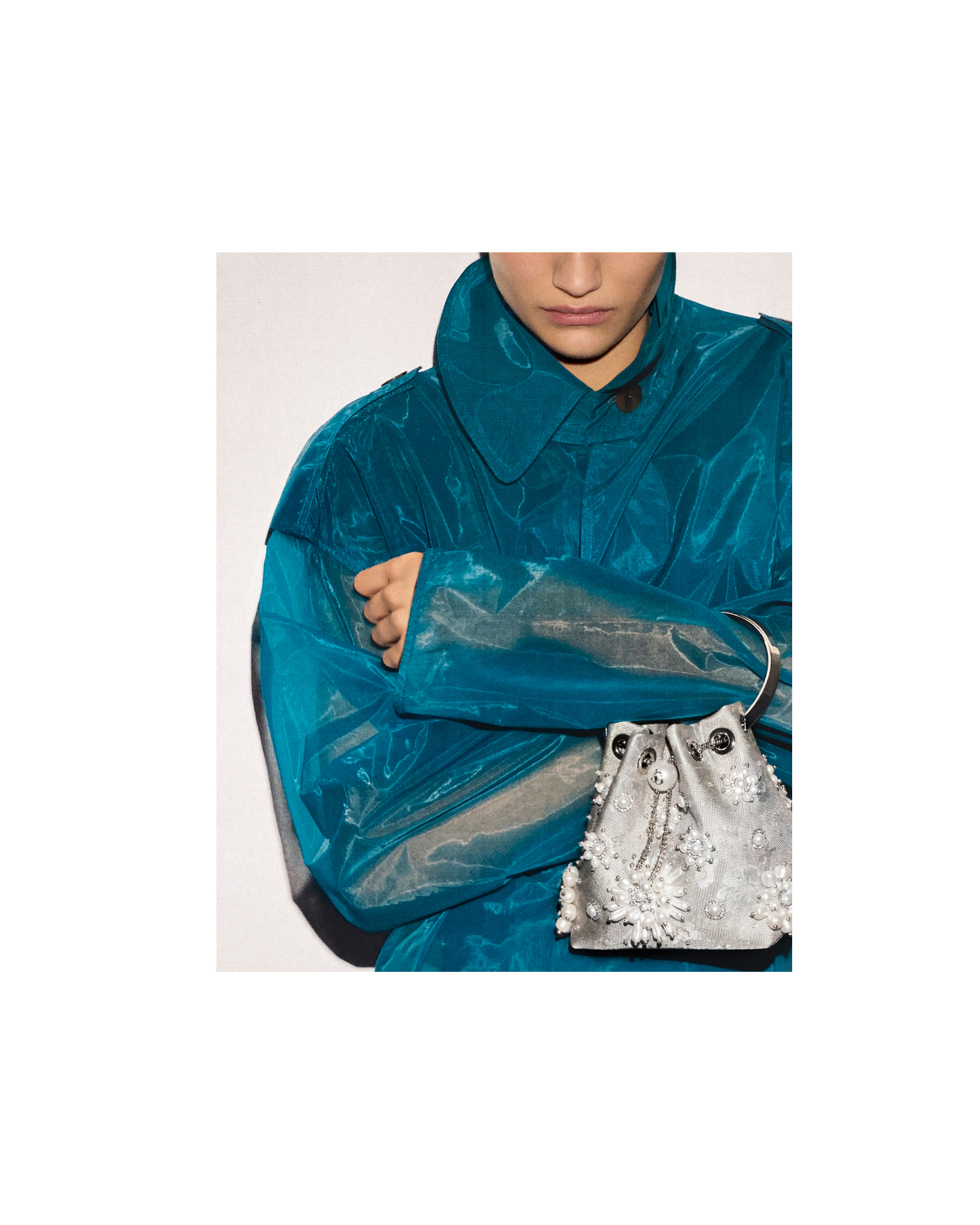 Model wearing a turquoise coat and holding a drawstring bag with pearl and crystal embellishments.
