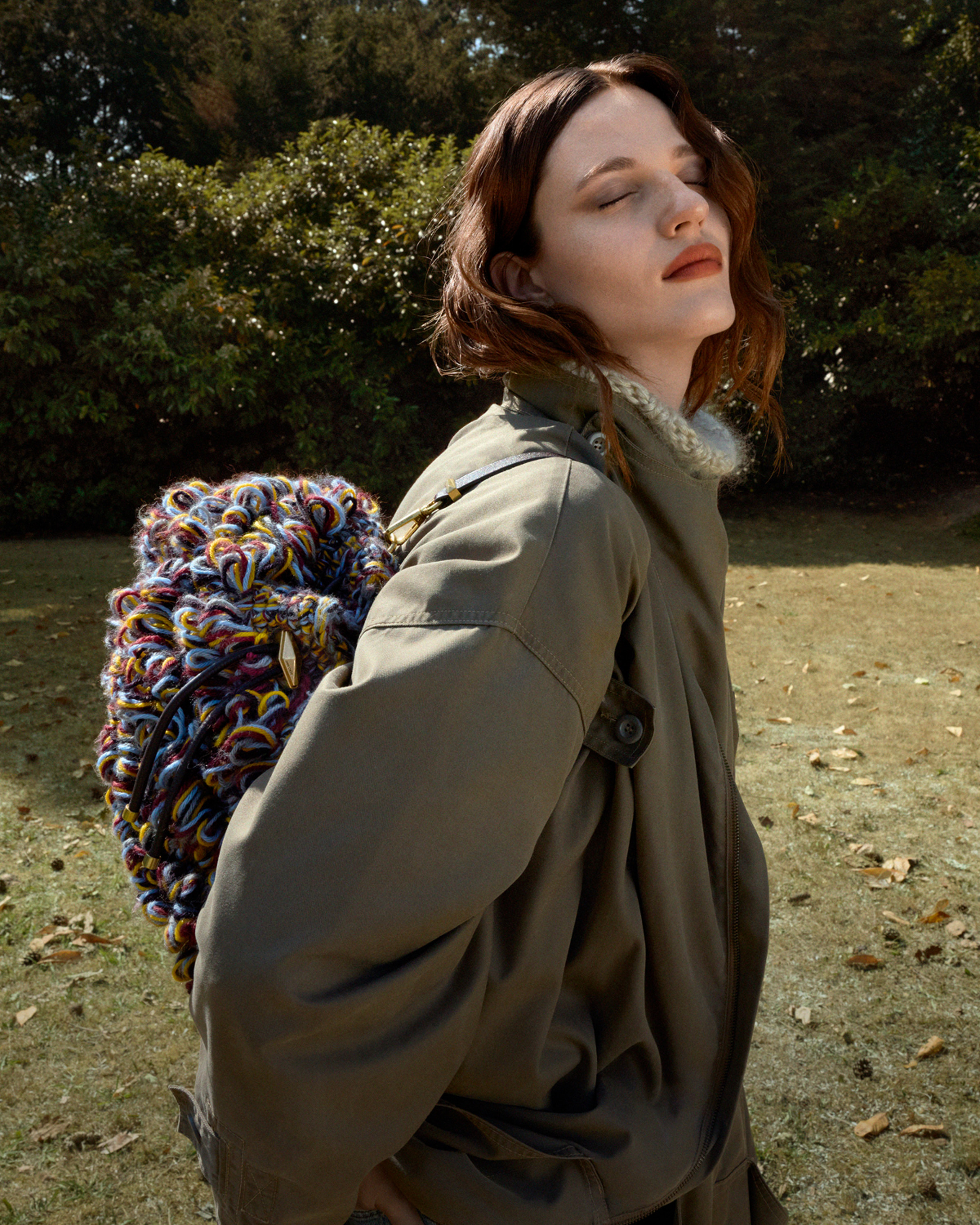 Model wearing a long khaki coat with a multicoloured woven knit bag carried on her back, standing outdoors on a lawn.