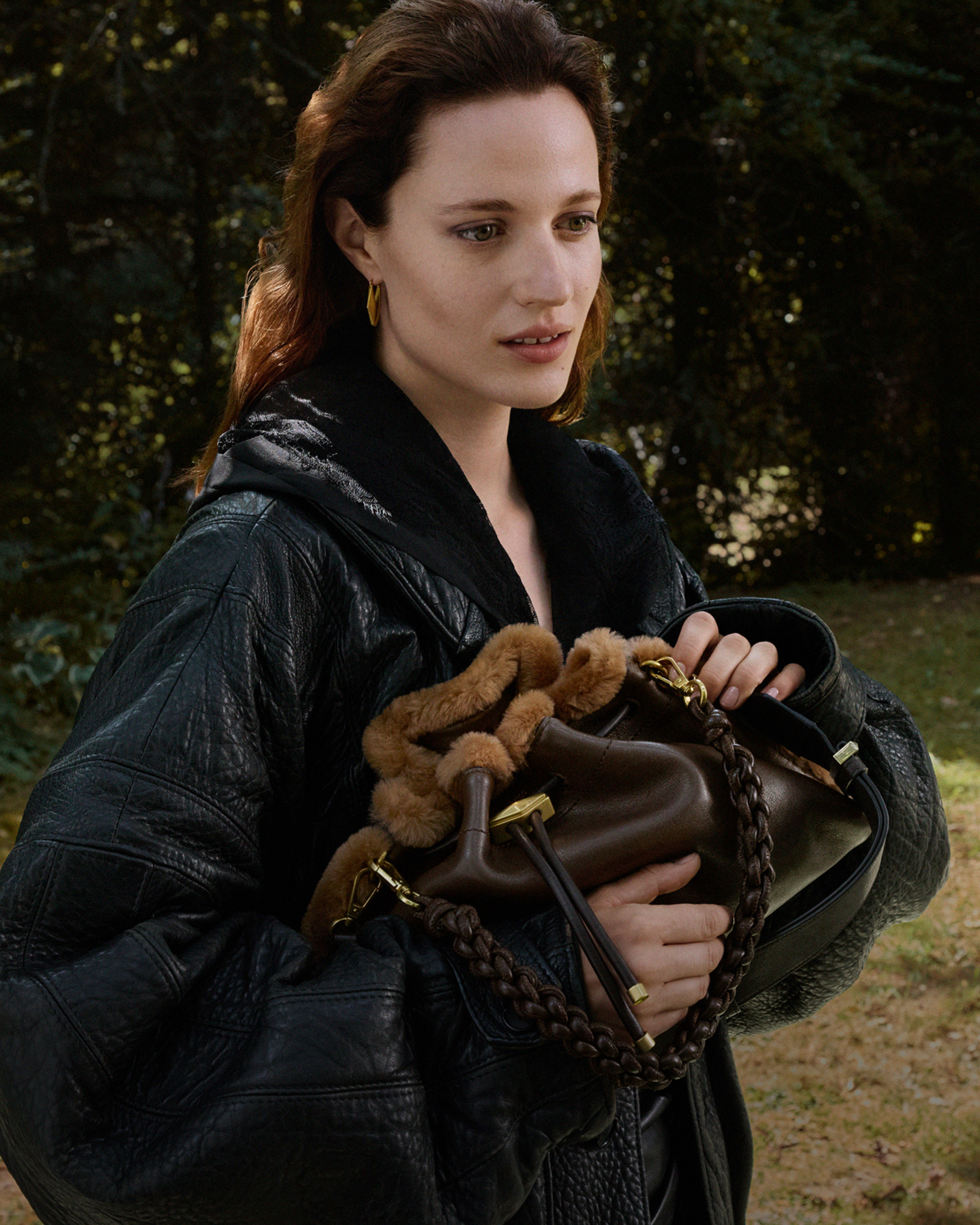 Model wearing a black leather jacket and holding a brown leather drawstring bag with shearling trim, braided handle and gold-tone hardware outdoors.
