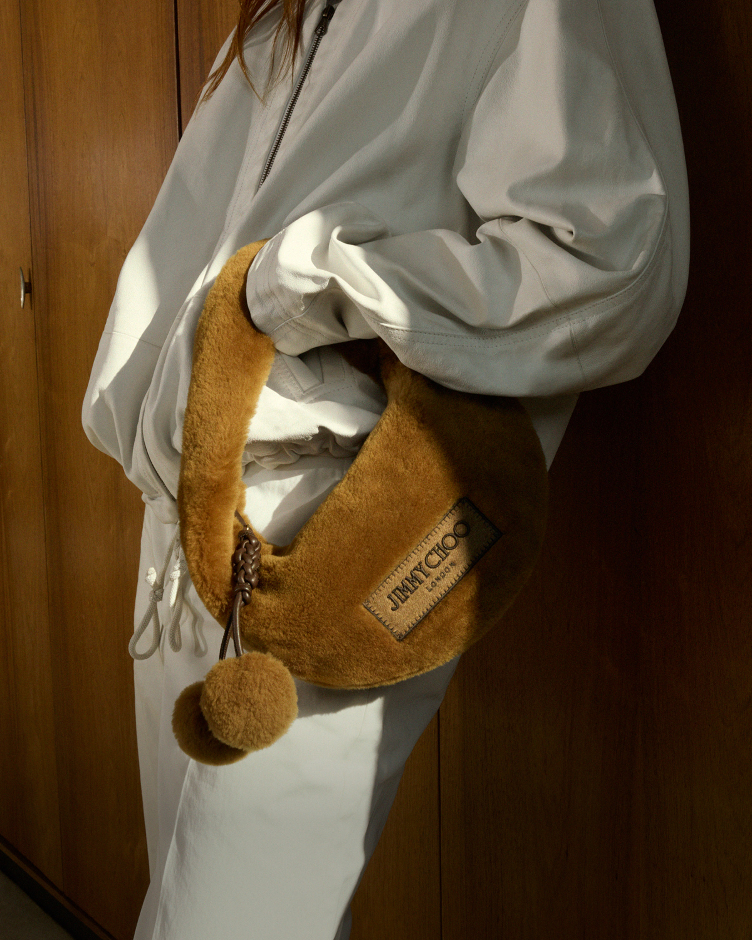  Close-up of a model wearing a cream outfit and carrying a brown shearling shoulder bag with a pom-pom detail.