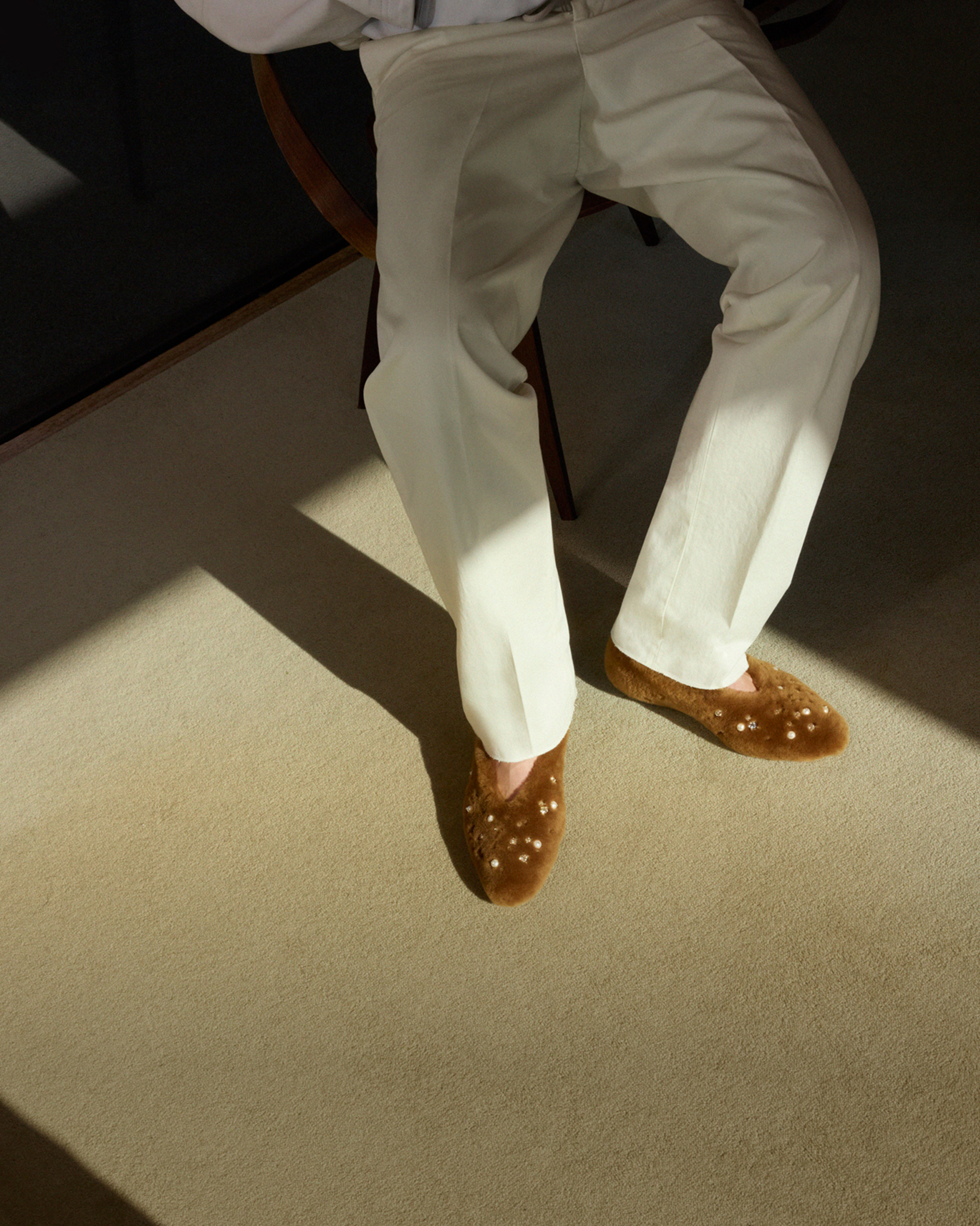 Close-up of brown shearling ballet flats with crystal and pearl embellishments, worn with cream trousers in a sunlit interior.