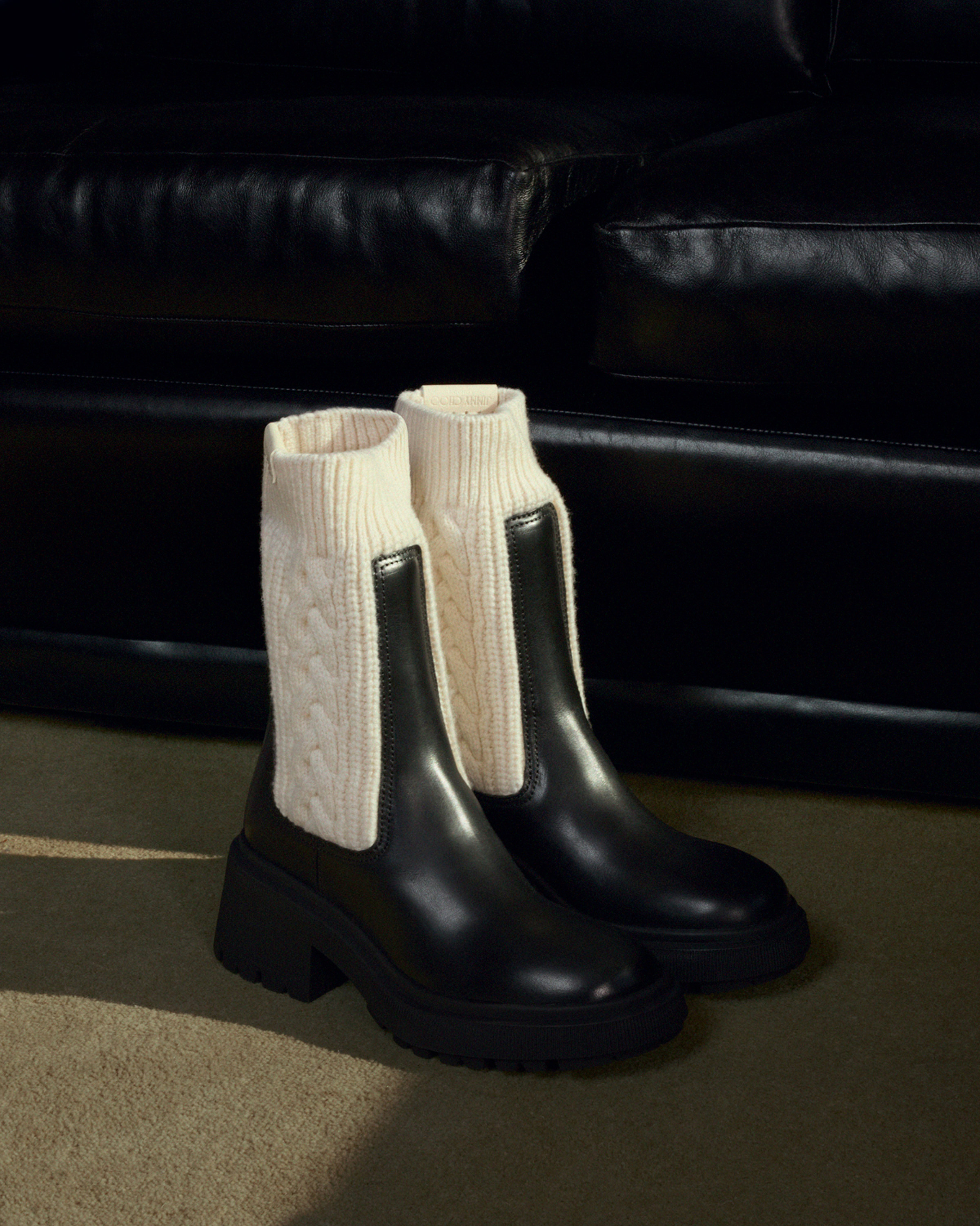  Pair of black leather Chelsea boots with cream cable-knit panels and chunky lug soles, displayed in front of a black leather sofa.