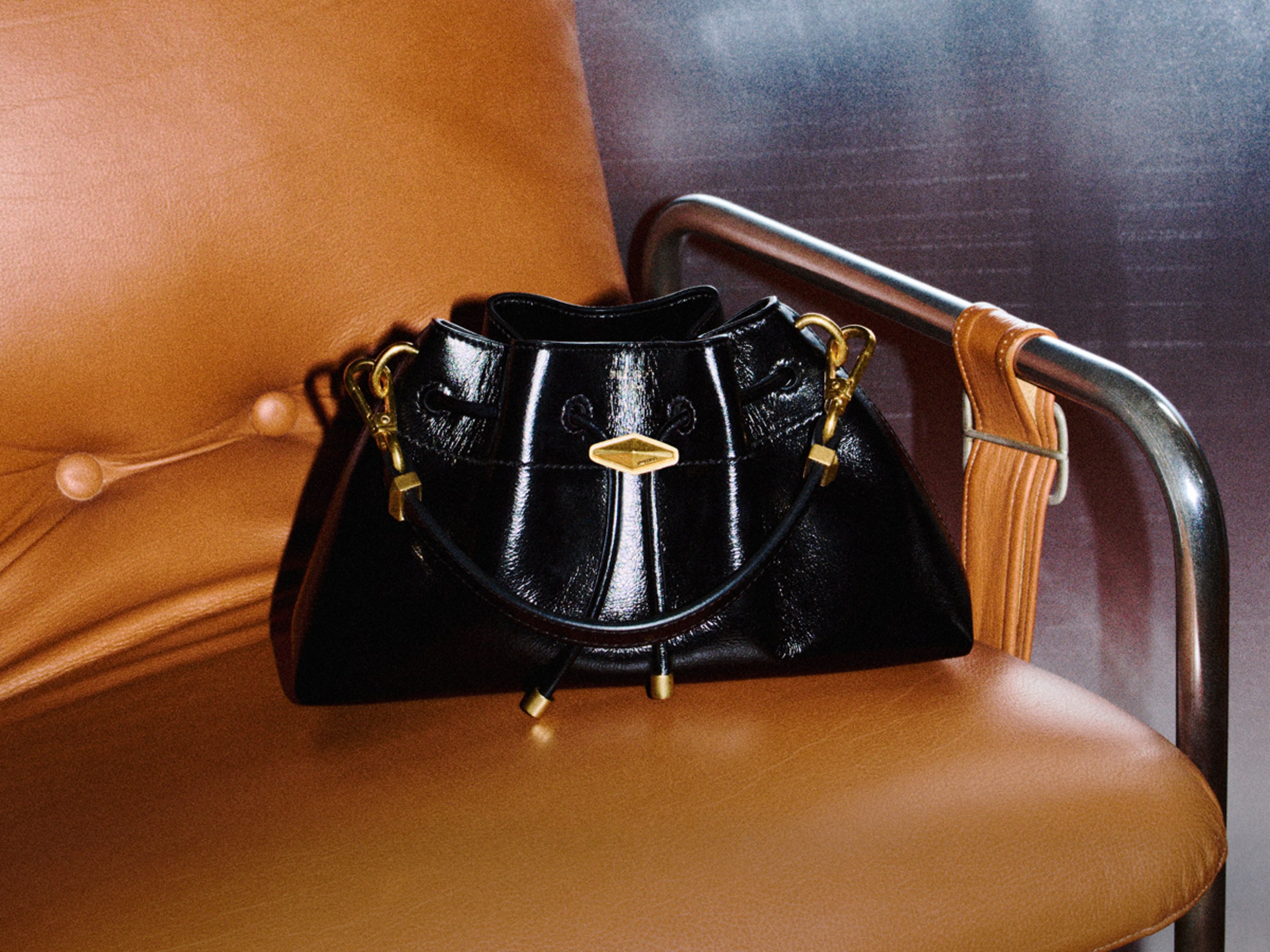 Black patent leather drawstring bag with gold-tone hardware displayed on a tan leather chair.