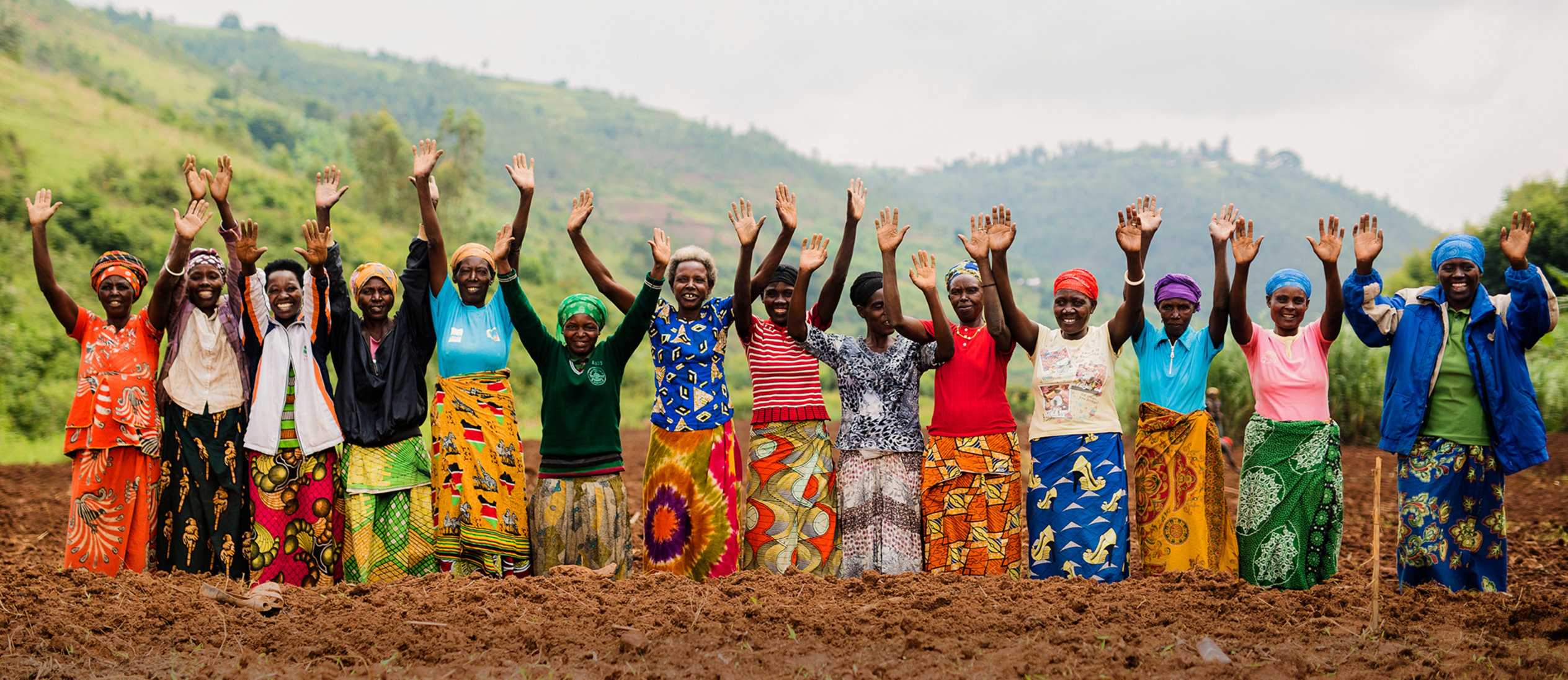 Group of women standing in a cultivated field with their hands raised, against a backdrop of green hills.