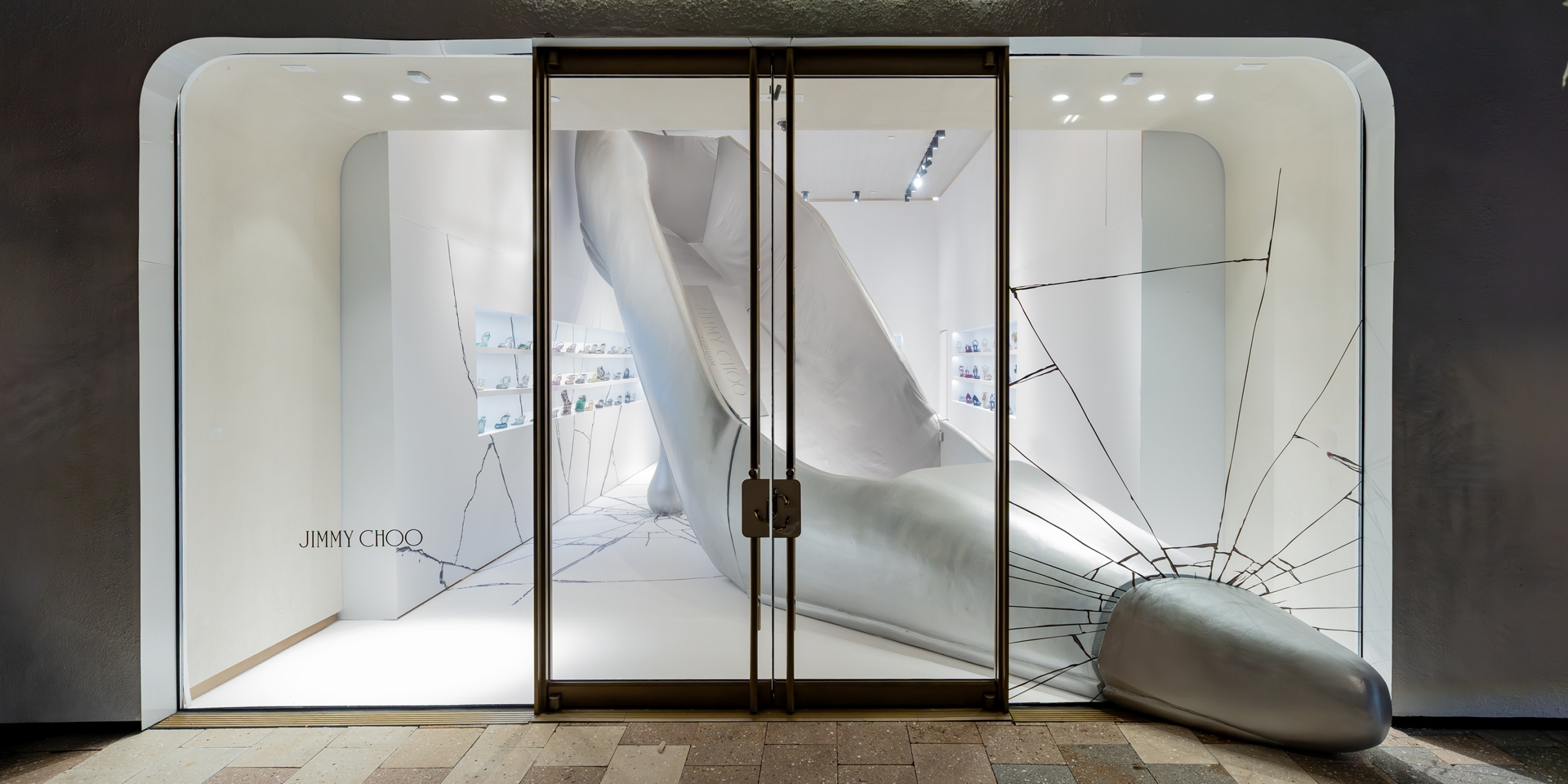Jimmy Choo store window featuring a giant sculptural silver high heel installation emerging through it with cracked floor detailing.