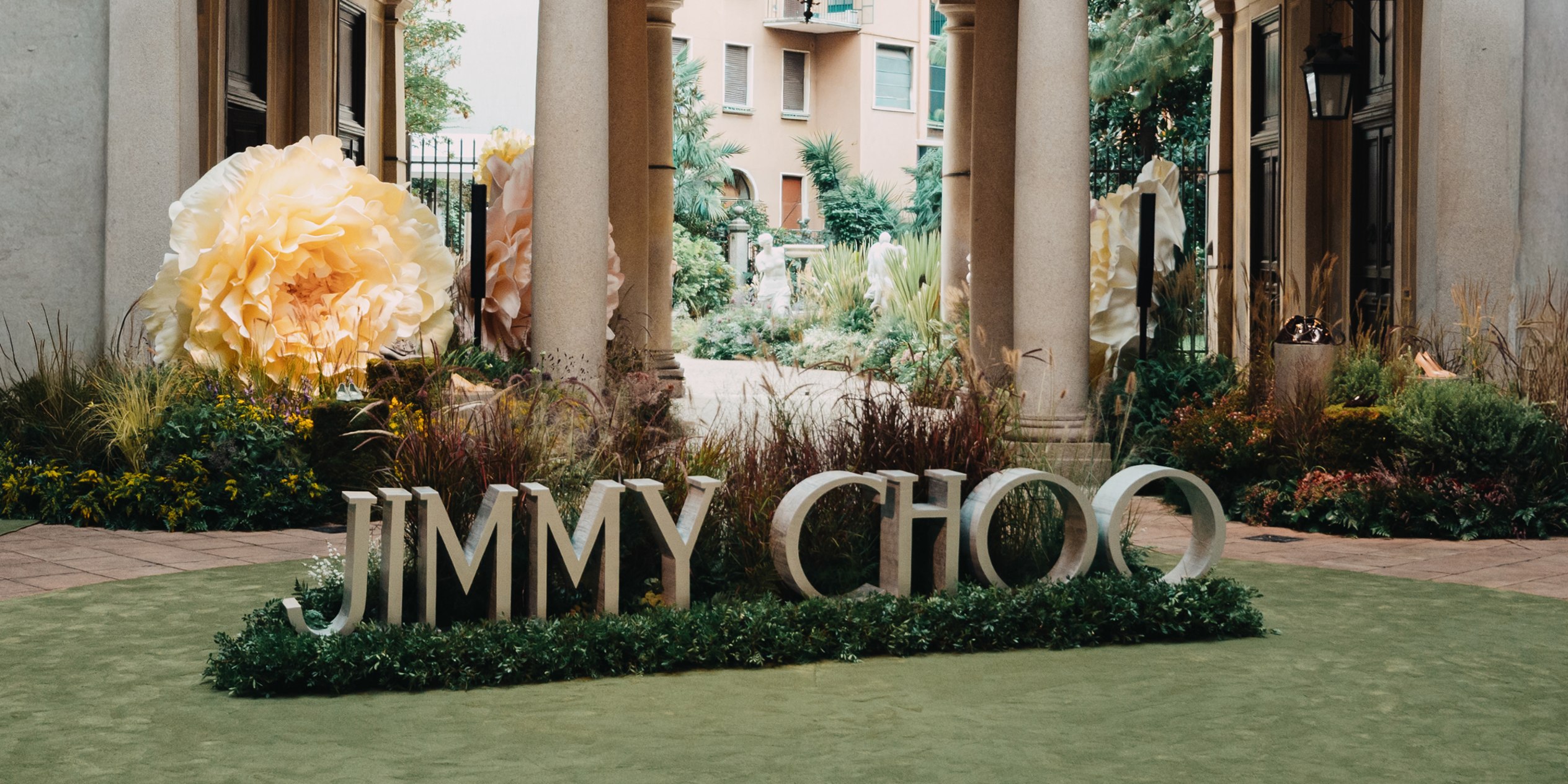 Close-up of Milan Fashion Week Jimmy Choo sign installation surrounded by greenery, with large pastel toned flowers amongst a garden backdrop and architectural pillars.