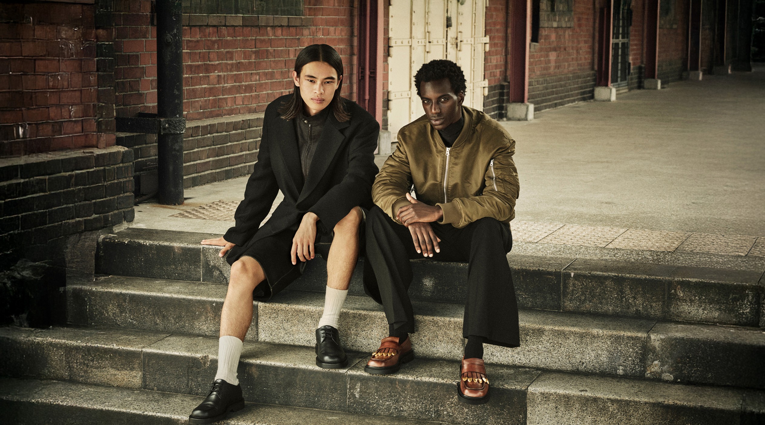 Two male models seated on stone steps wearing black and brown outerwear with loafers and sandals.			