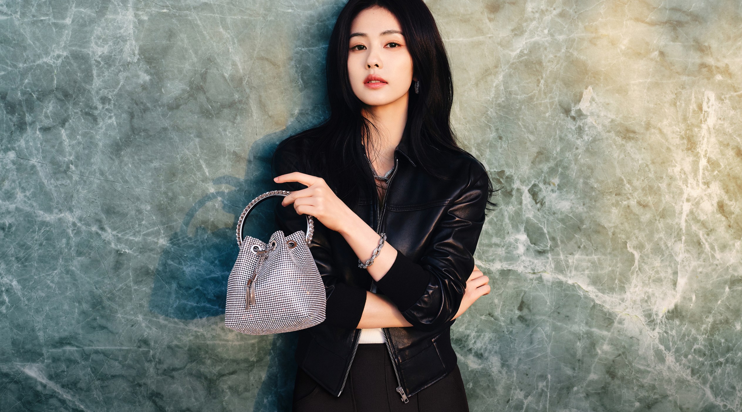 Woman in black leather jacket holding crystal-embellished clutch against a textured