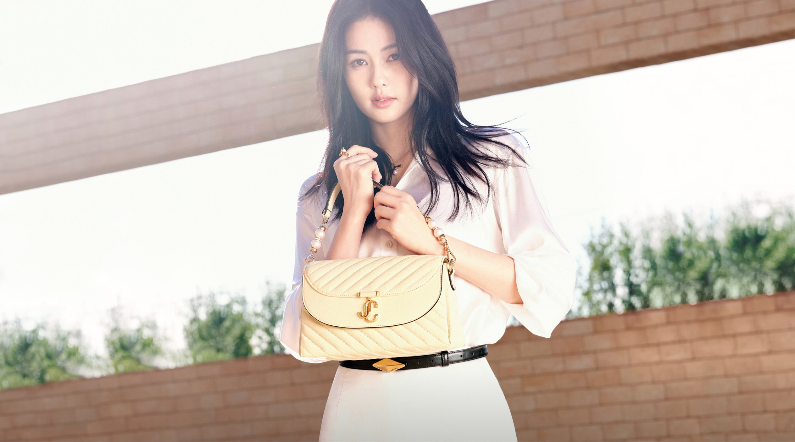 Woman sitting on a white bed in a neutral room, wearing black woven heels and holding a raffia Jimmy Choo tote bag with black leather trim and drawstring closure.