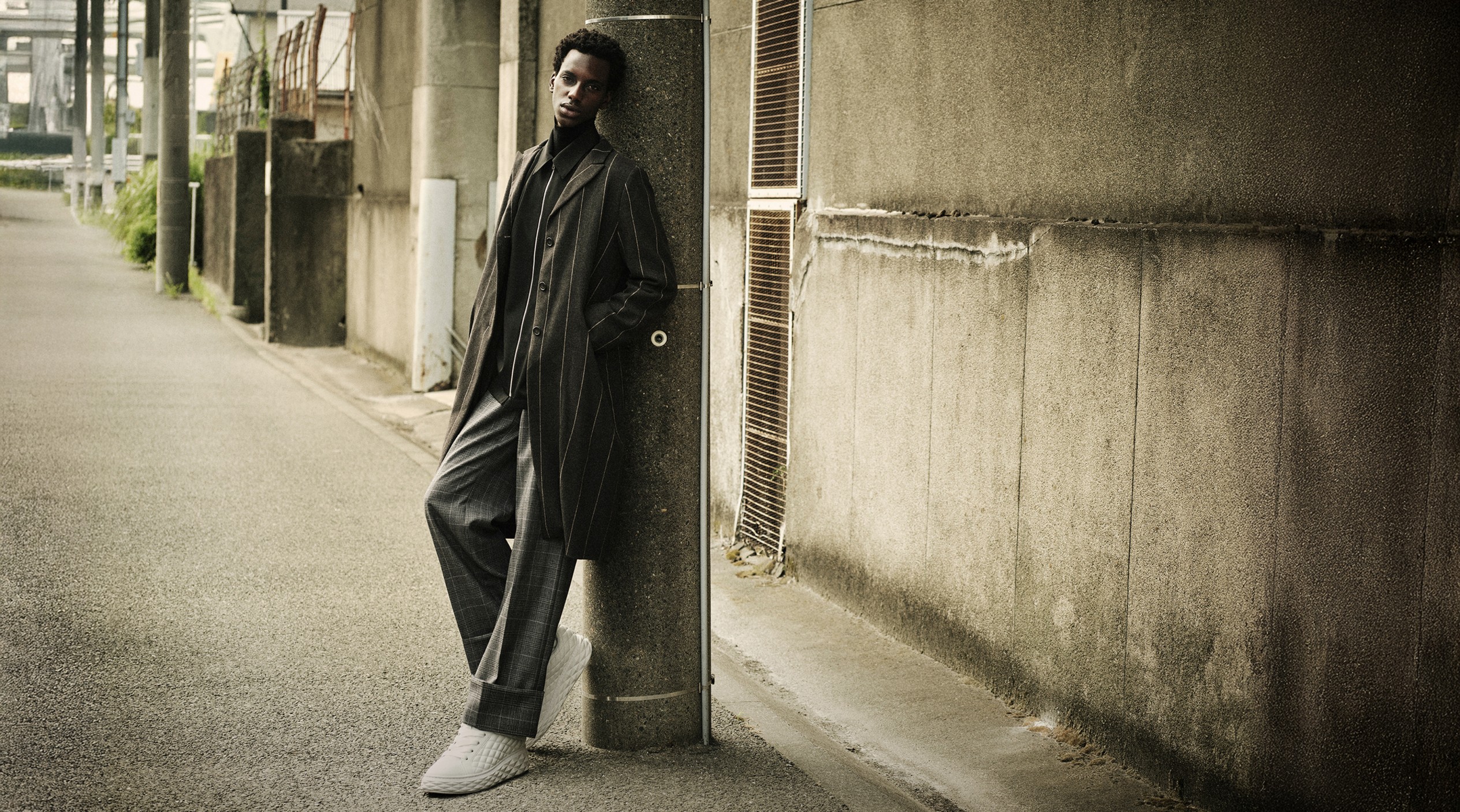 Man leaning against a concrete post in an urban street, wearing a lomg dark coat, striped trousers, and white sneakers.