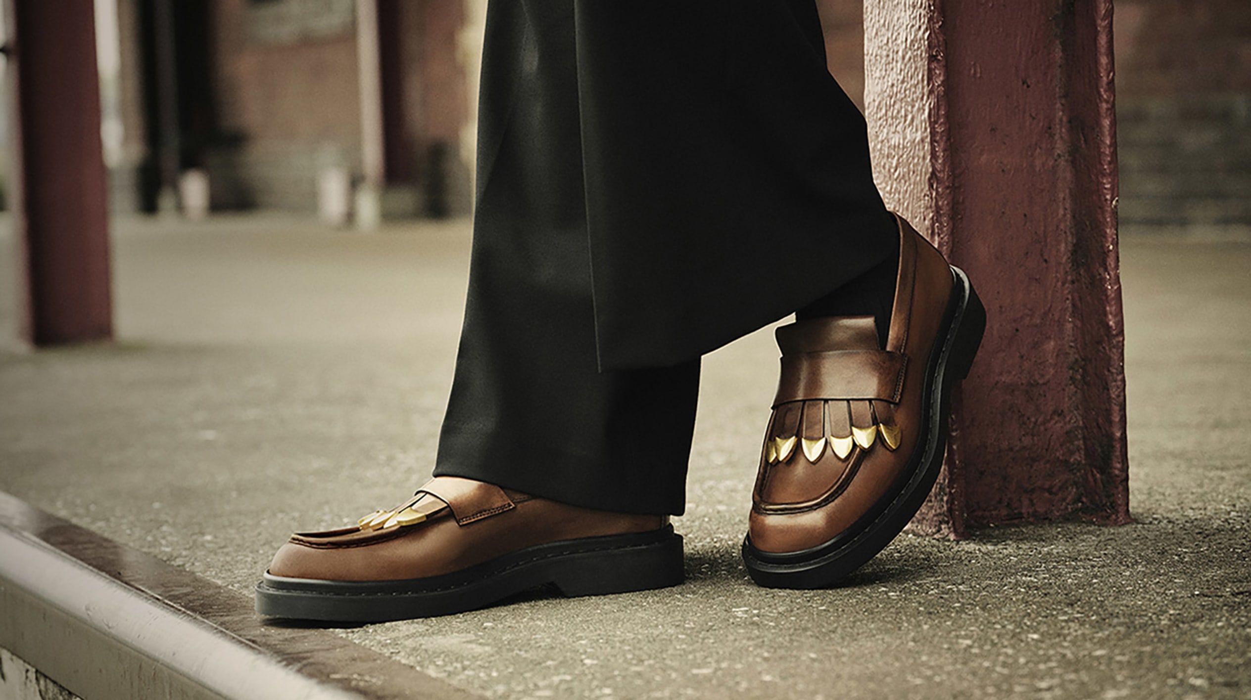 Man wearing brown leather loafers with fringe detail, styled with black trousers, standing against a brick building backdrop.