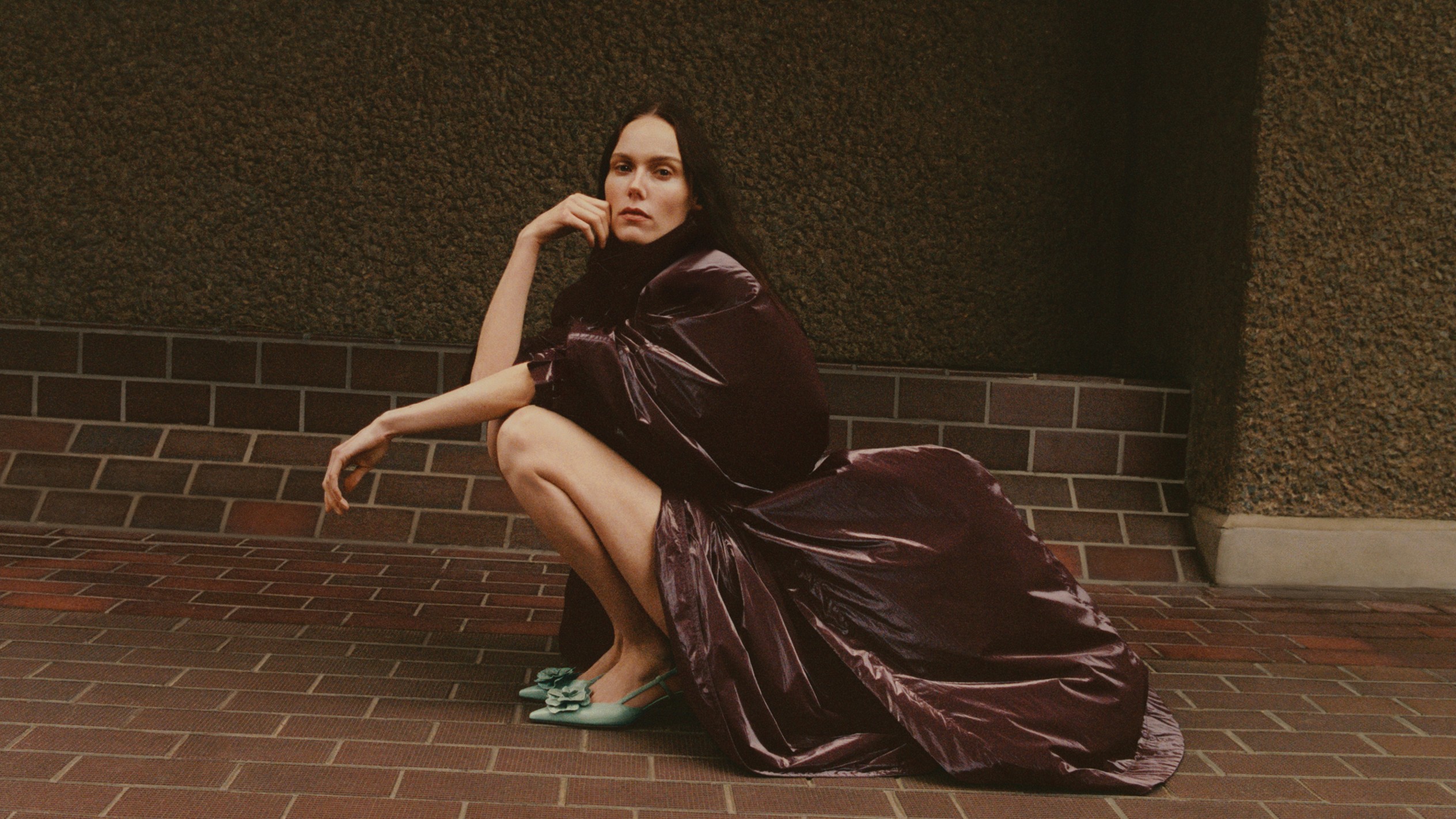 Woman crouching on a brick pavement wearing a dark, glossy dress and pale green pointed slingback heels with floral corsage accent, against a textured wall.