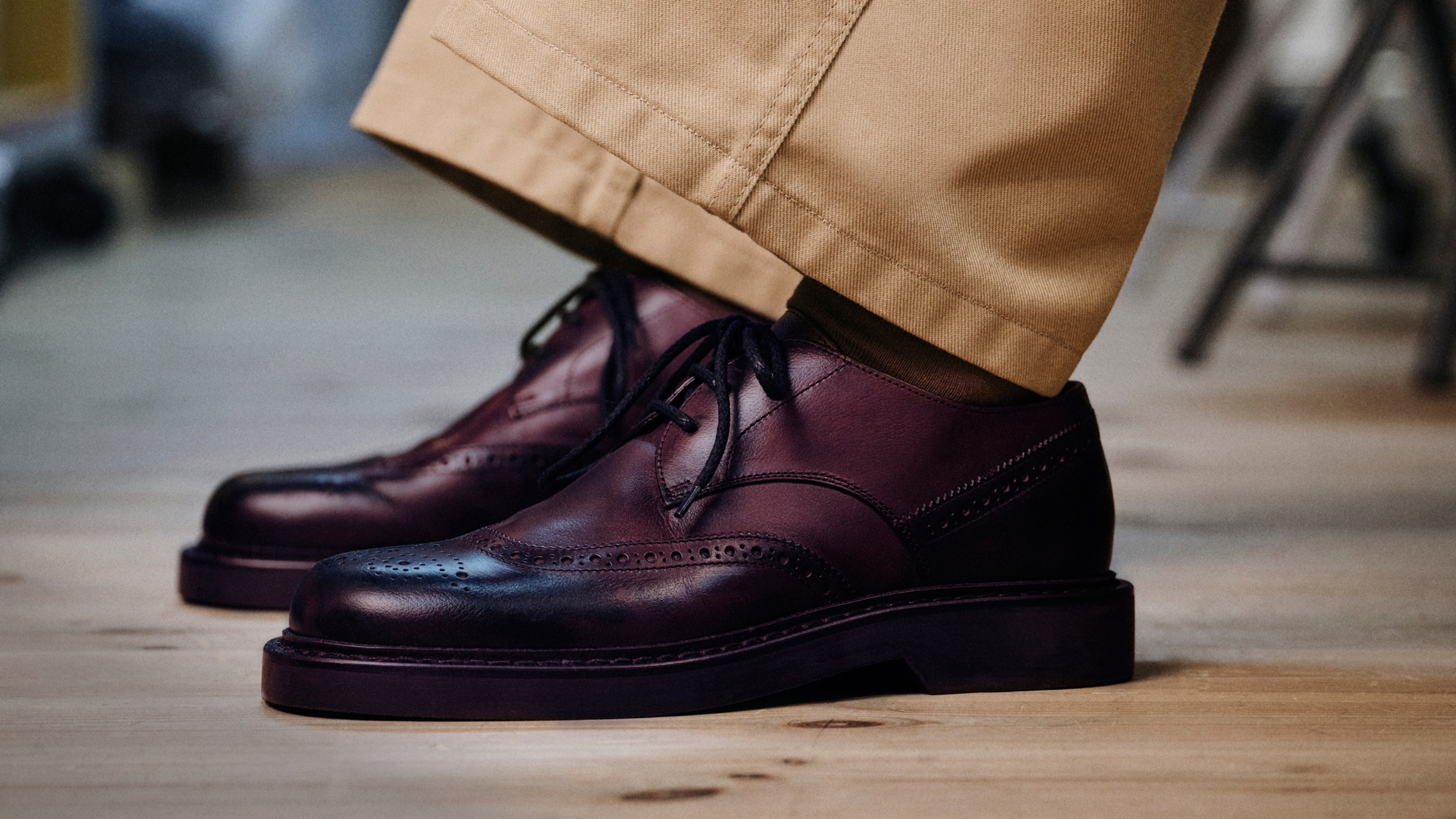 Burgundy leather lace-up shoes worn with cropped beige trousers on a wooden floor.