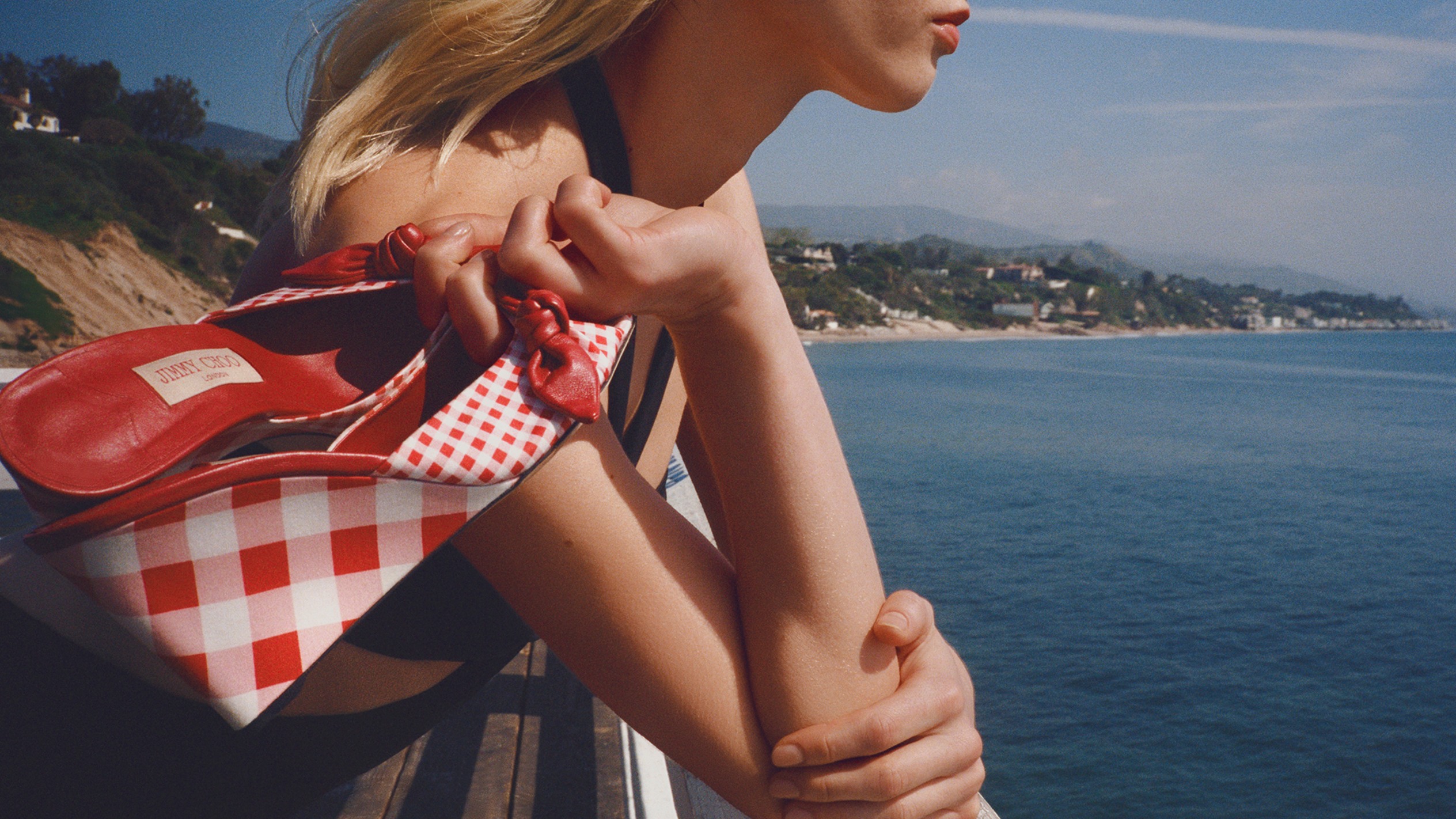 Model walking between tall hedges at sunset carrying a blue gingham shoulder bag and wearing a beige trench coat.			