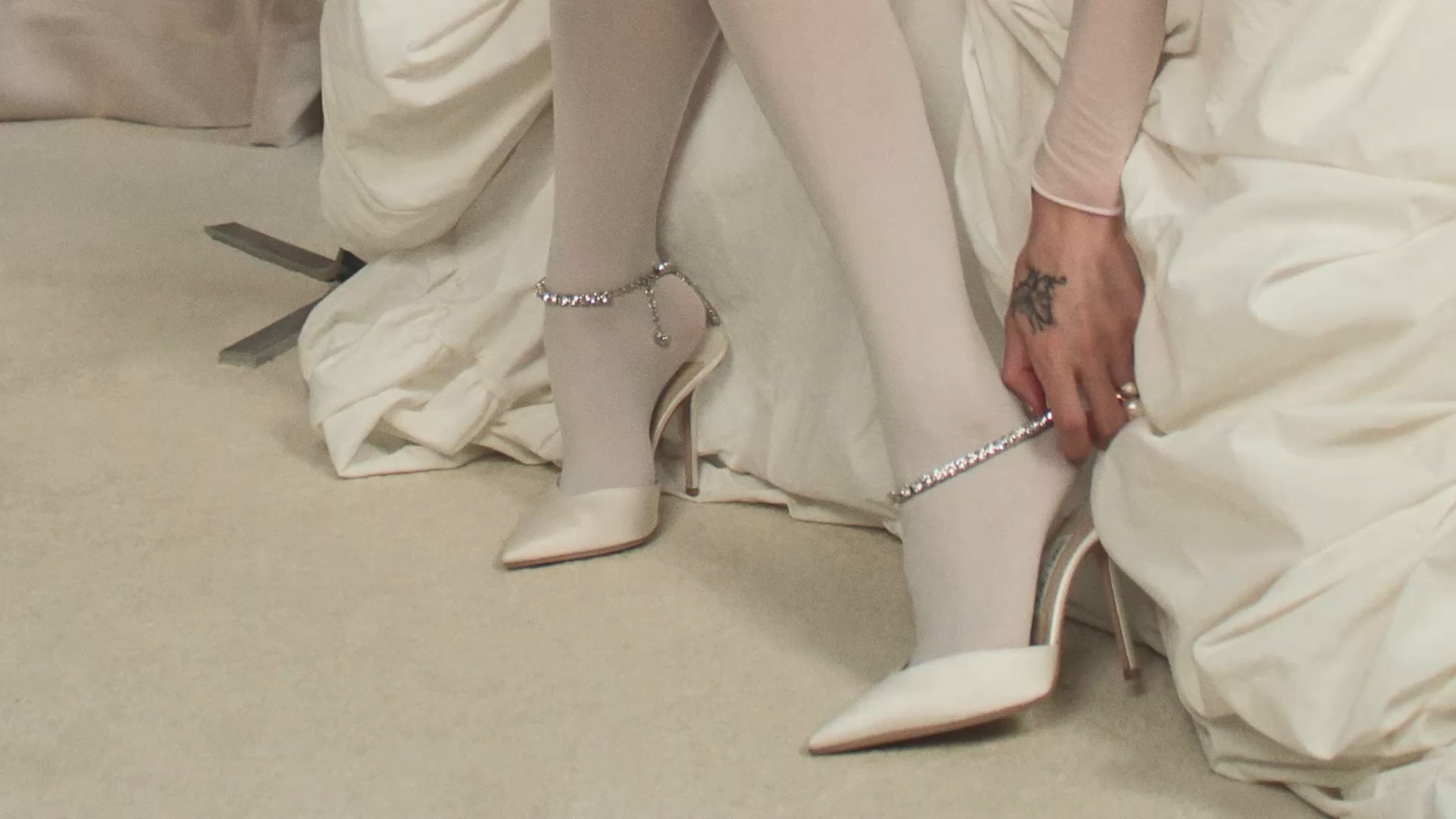 Model wearing pearl-embellished pointed-toe heels while seated on a chrome chair against a blue backdrop.