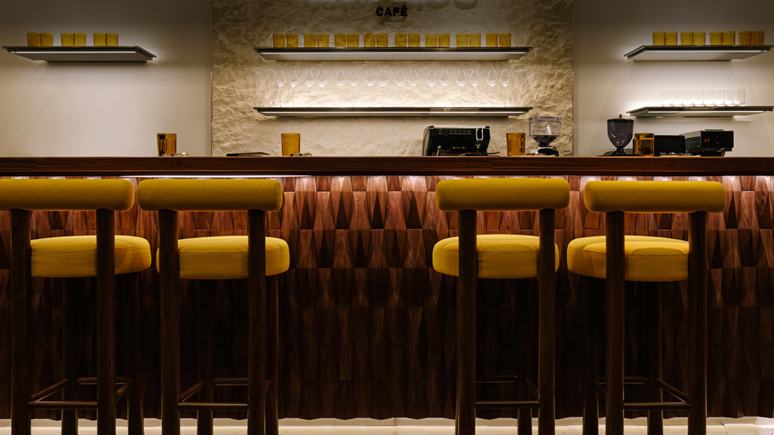 柔らかい照明が当たる棚と木製パネル、黄色いバースツールが並ぶ Jimmy Choo Caf&eacute; のバー カウンター。 