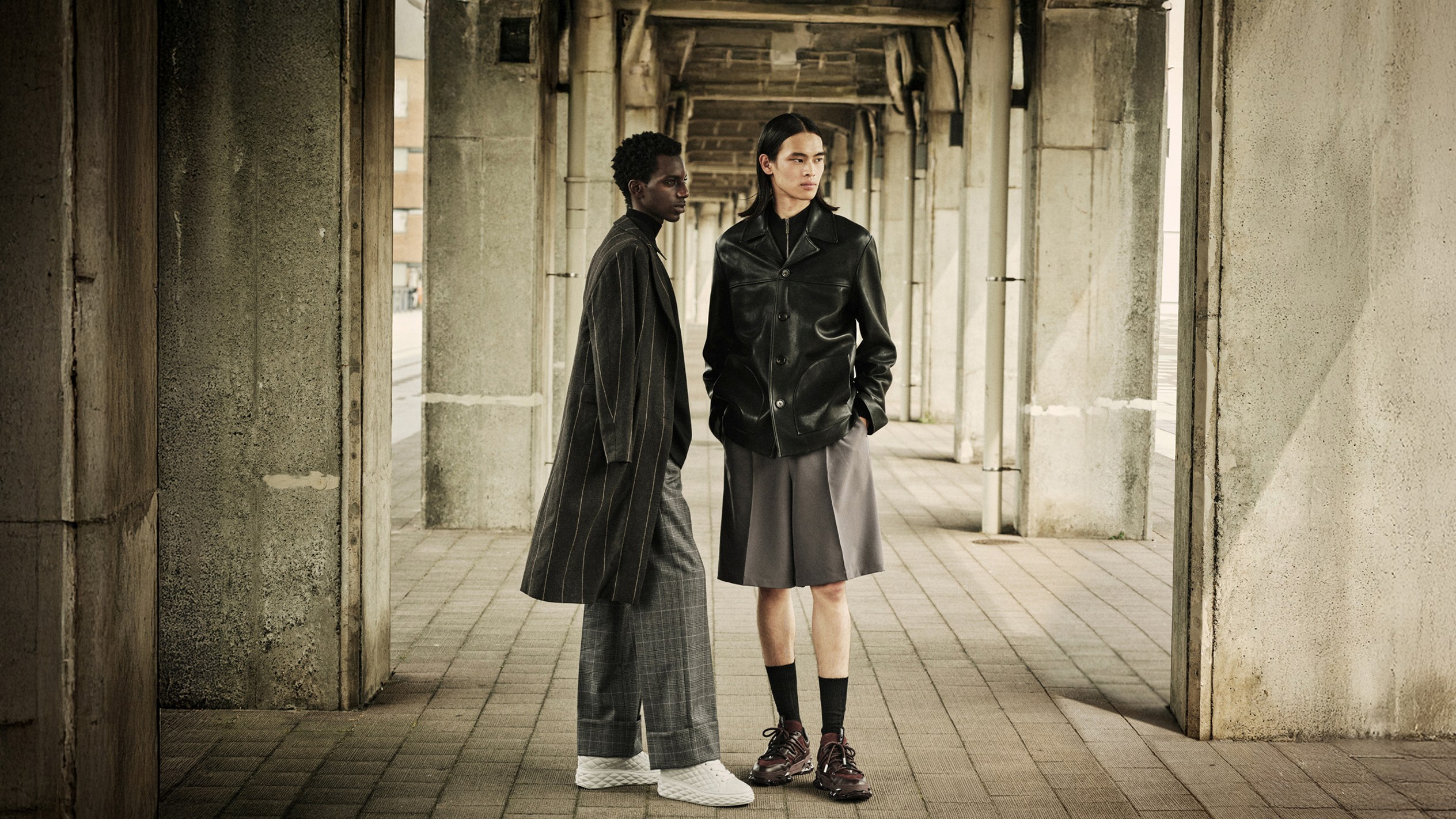 Two male models in layered autumn looks, one in a long pinstripe coat with wide-leg trousers and trainers, the other in a leather jacket styled with tailored shorts and loafers, shot in an architectural walkway.