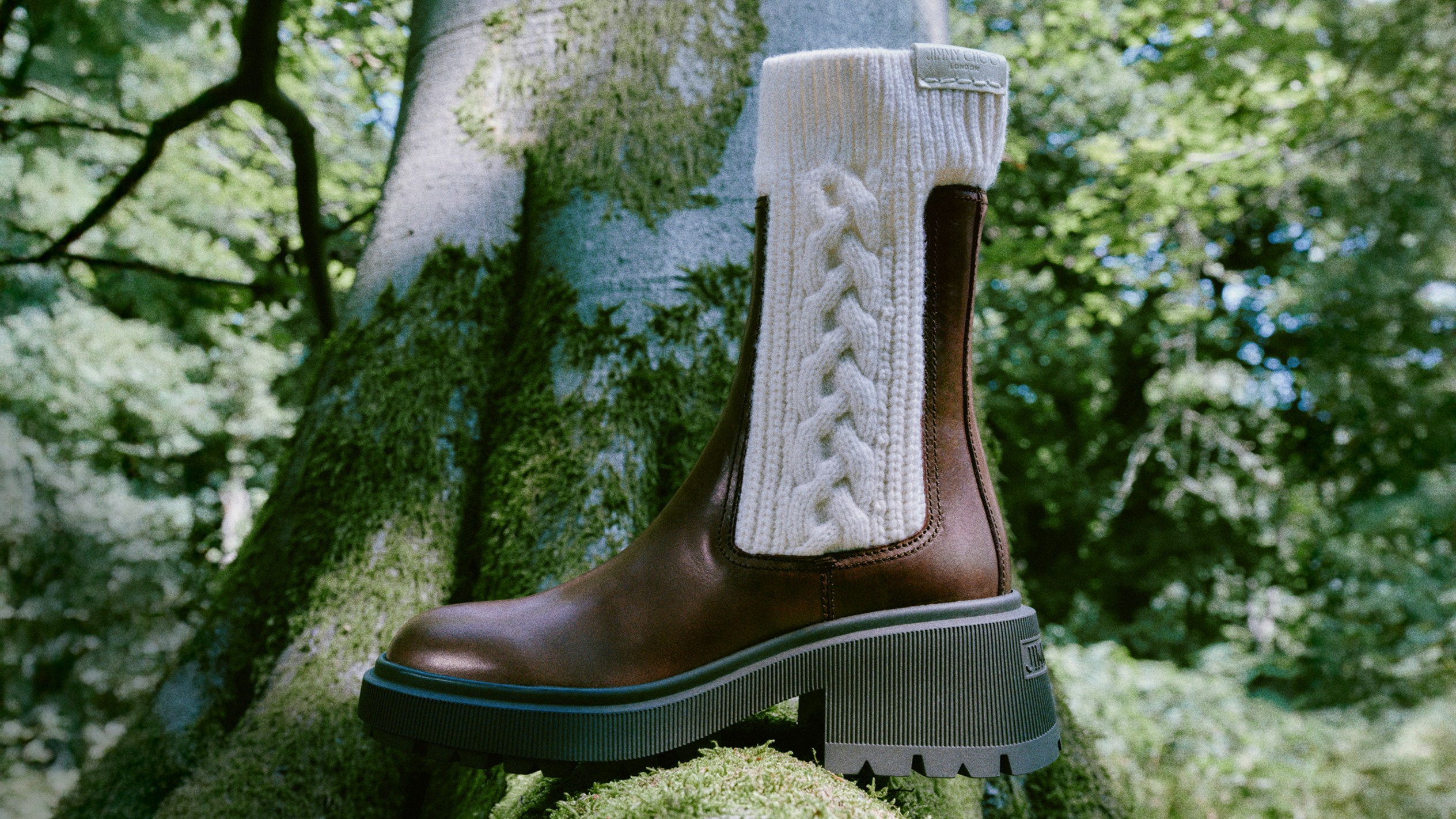 Brown leather ankle boot with chunky sole and cream cable-knit panel, photographed outdoors against a tree trunk in a forest setting.			