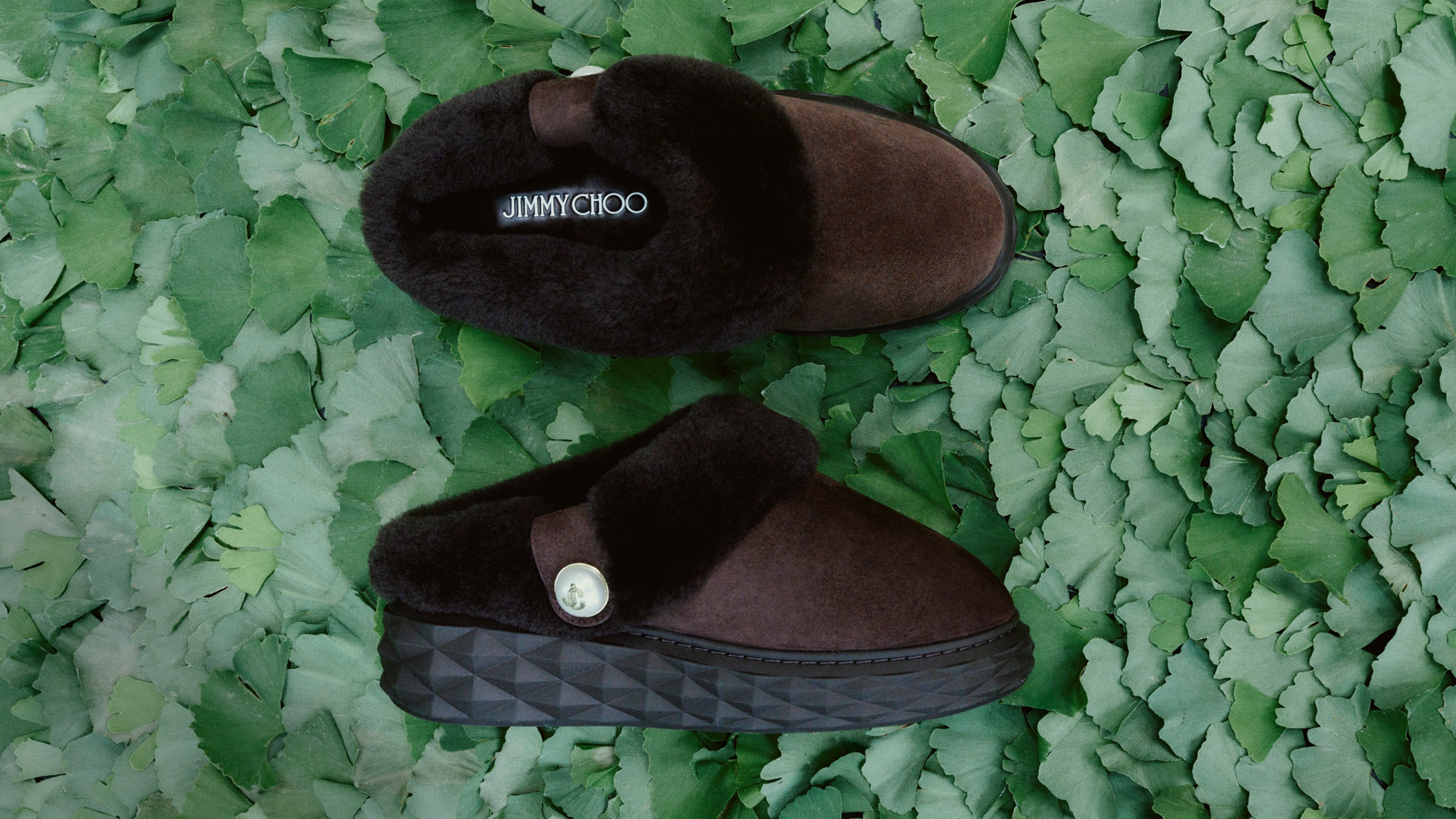 Brown shearling-lined slip-on mules with a chunky textured sole, photographed on a bed of green leaves.