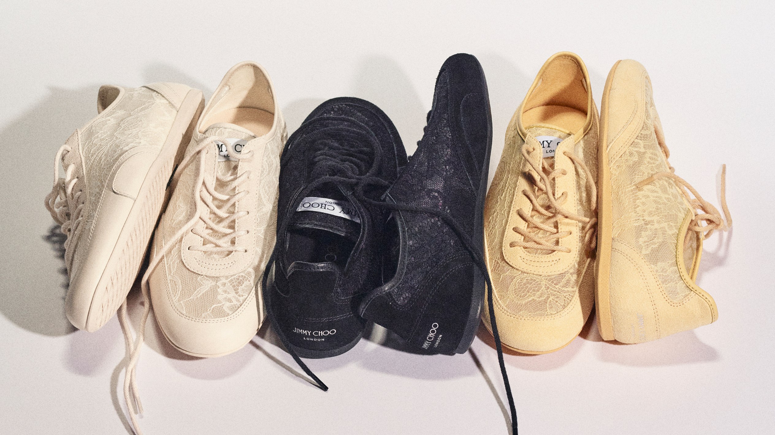 A lineup of Jimmy Choo Sunny trainers arranged side-by-side on a light surface, featuring lace-detailed uppers in latte, black, and yellow tones.