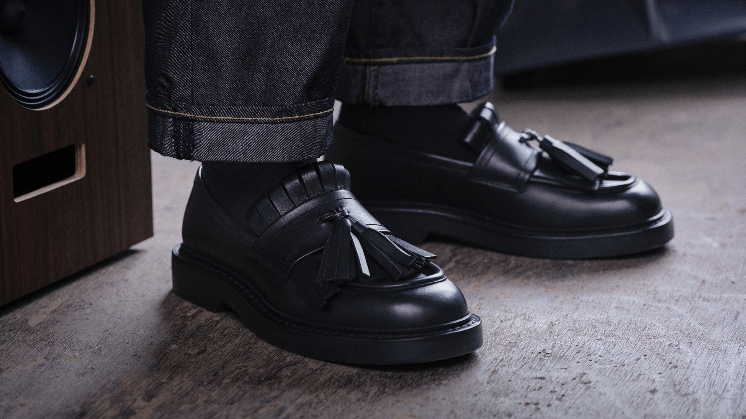 Black leather men&rsquo;s loafers with tassel detailing, worn with dark denim trousers, photographed on a concrete floor.