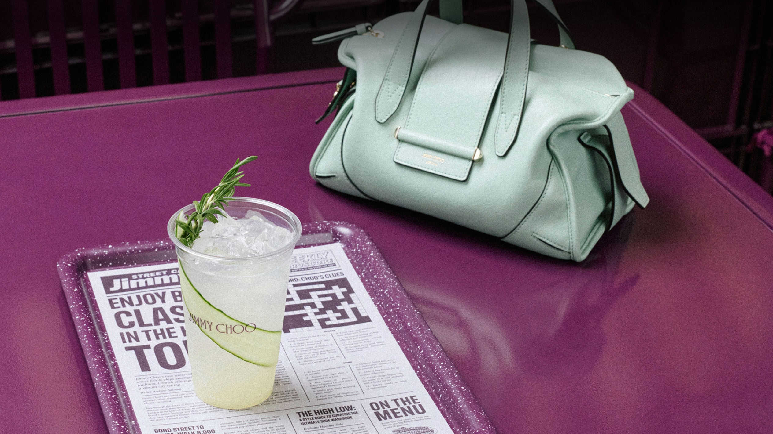 Light green holdall bag and iced drink on top of a Jimmy Choo newspaper on a purple caf&eacute; table.