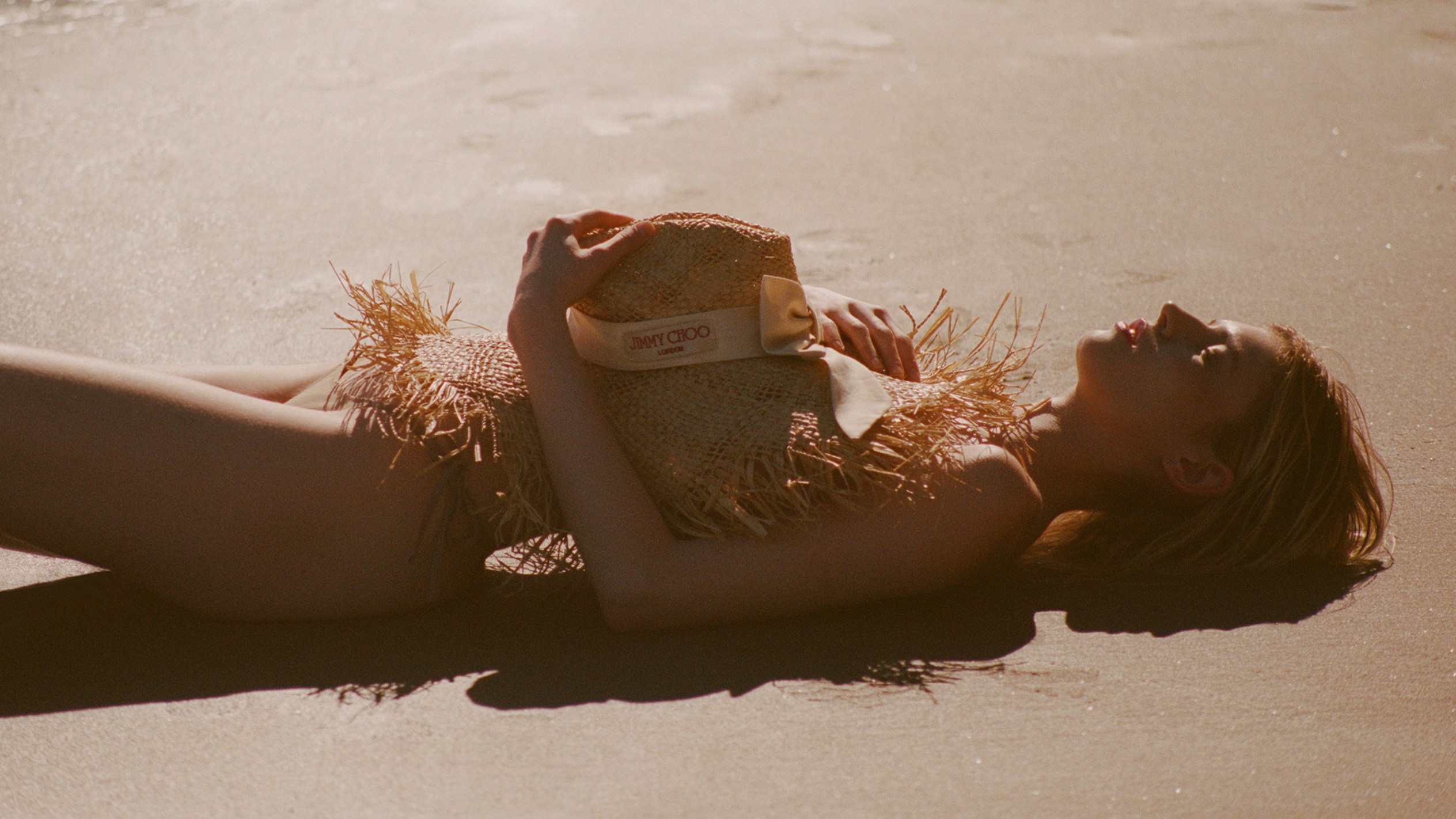 Model walking between tall hedges at sunset carrying a blue gingham shoulder bag and wearing a beige trench coat.			