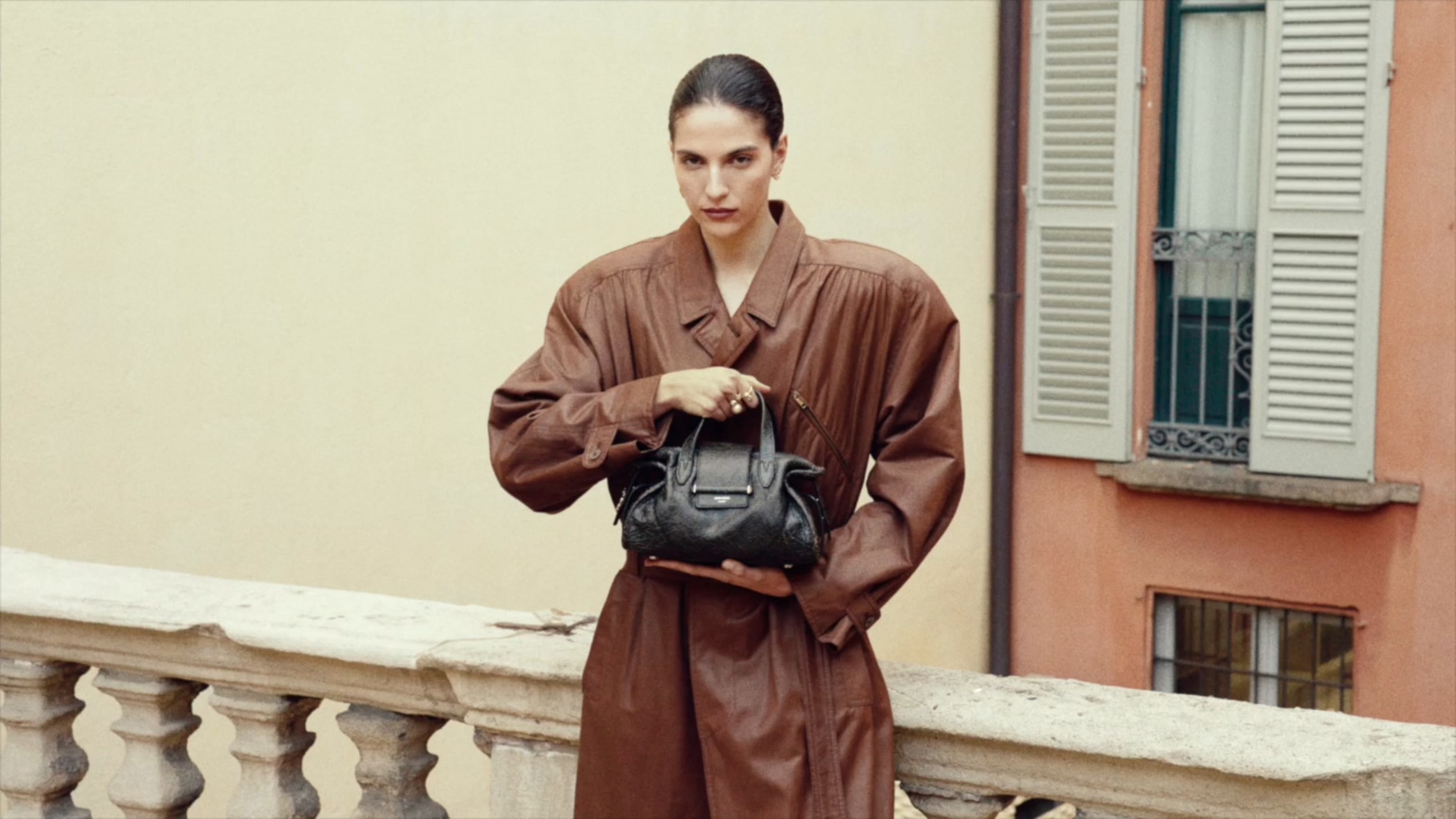 Model wearing a brown leather trench coat and holding a black leather top-handle bag while standing on a stone balcony.