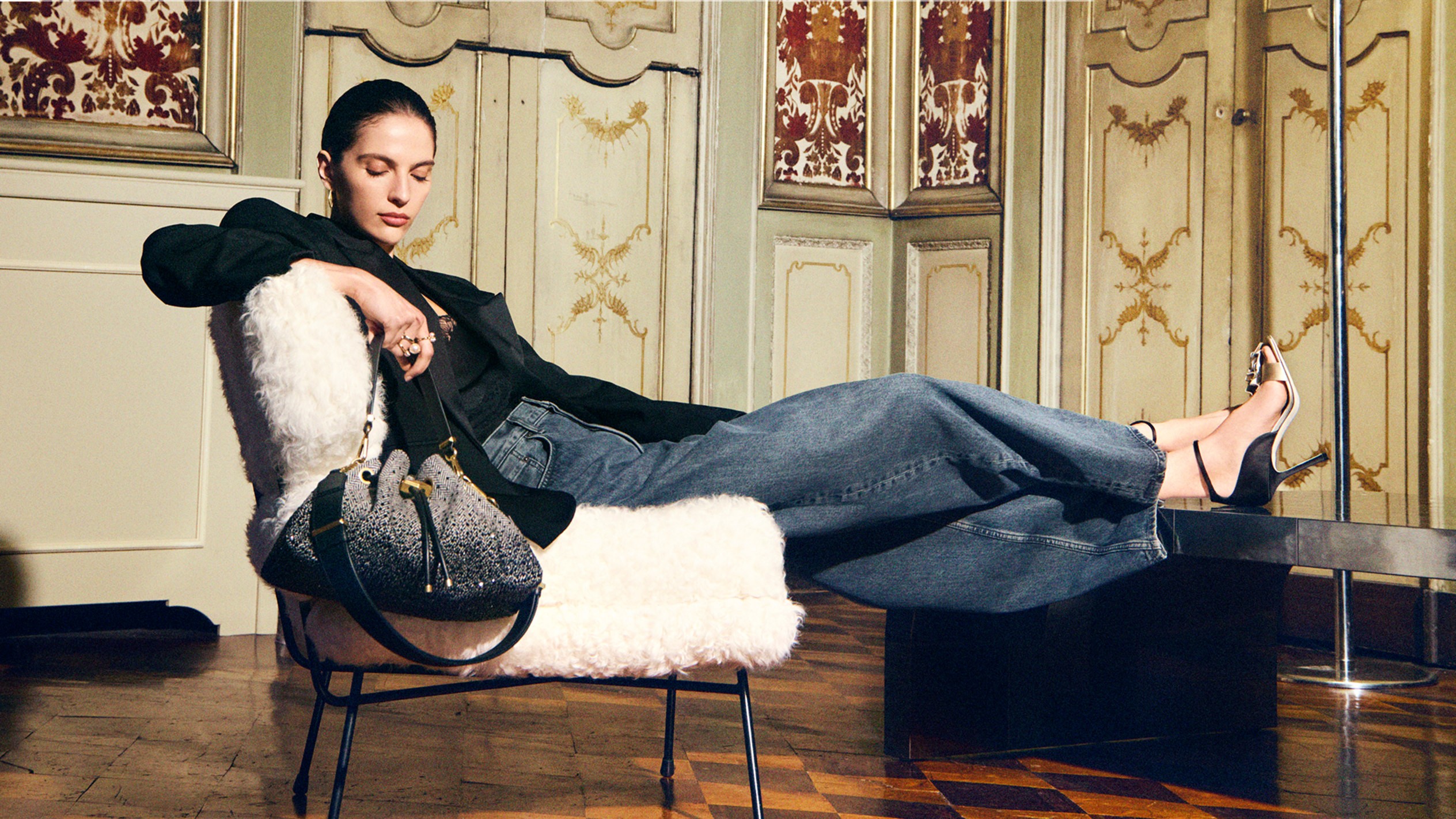 Model reclining on a shearling chair in an ornate interior, wearing a black top, wide-leg jeans and black slingback heels, with a crystal-embellished drawstring bag beside her.