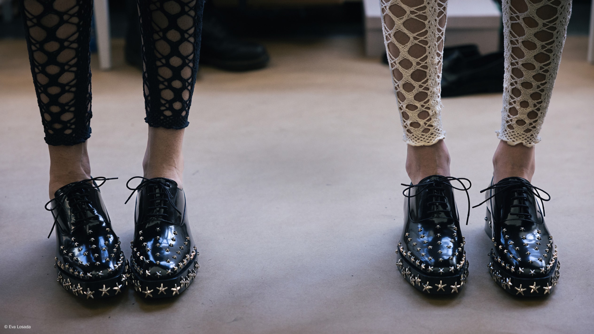 Two pairs of legs wearing black leather star studded lace-up shoes, styled with contrasting cut-out patterned leggings.