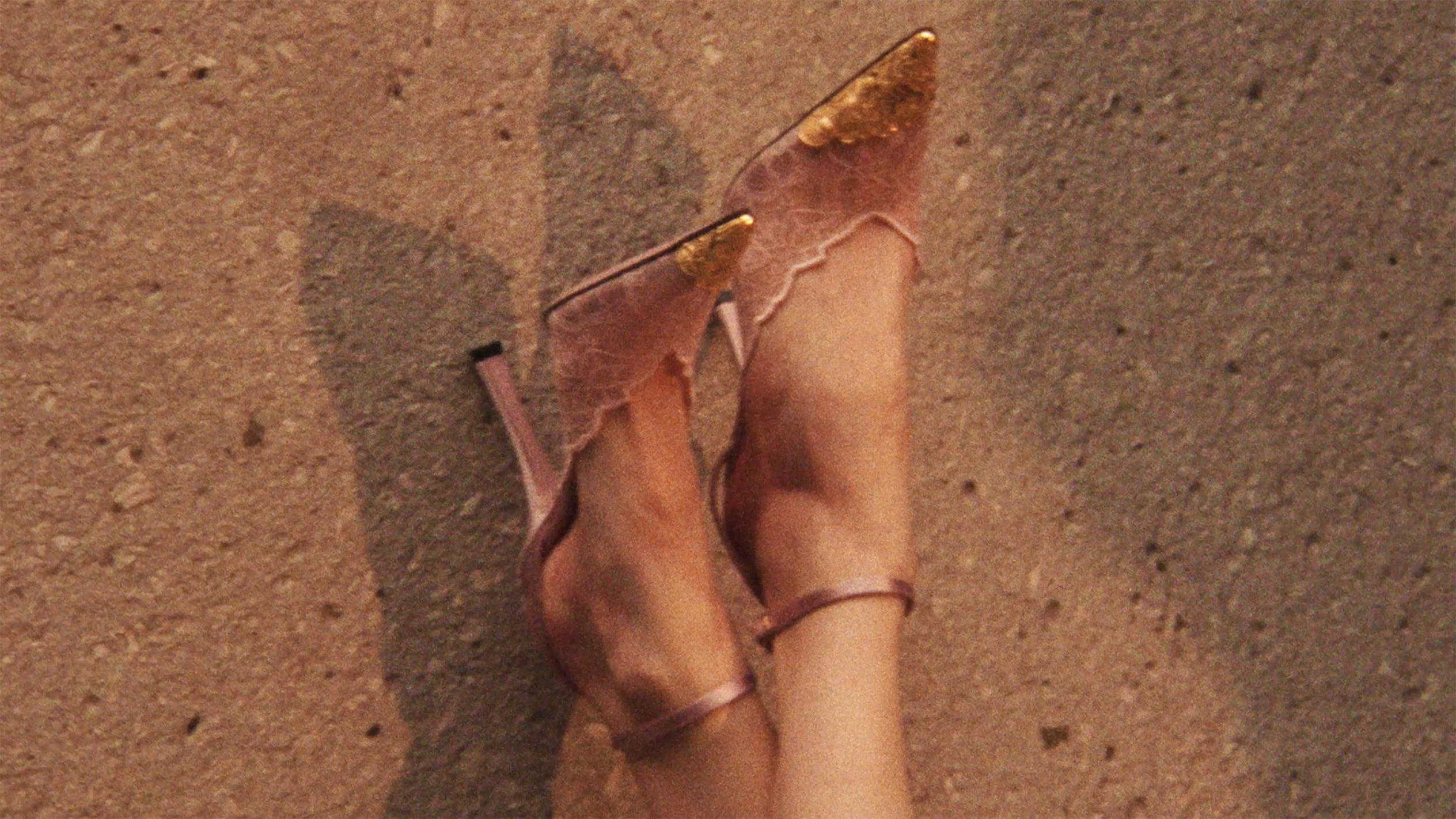 Sheer lace pointed-toe heels in soft pink lace, with metallic gold toe cap, styled against a textured stone surface.