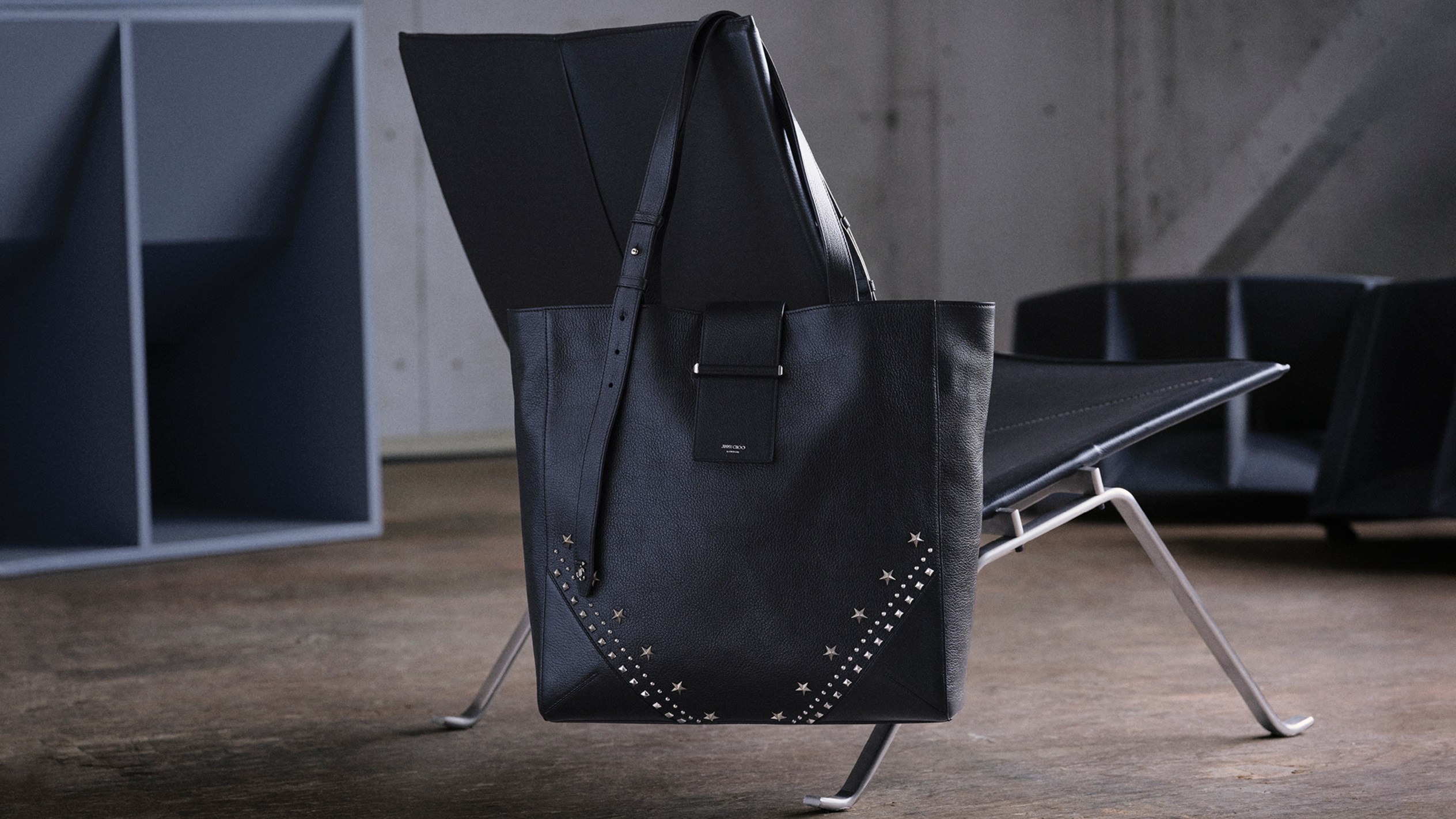 Black leather tote bag with silver star stud detailing, styled indoors hanging on a black modern chair.