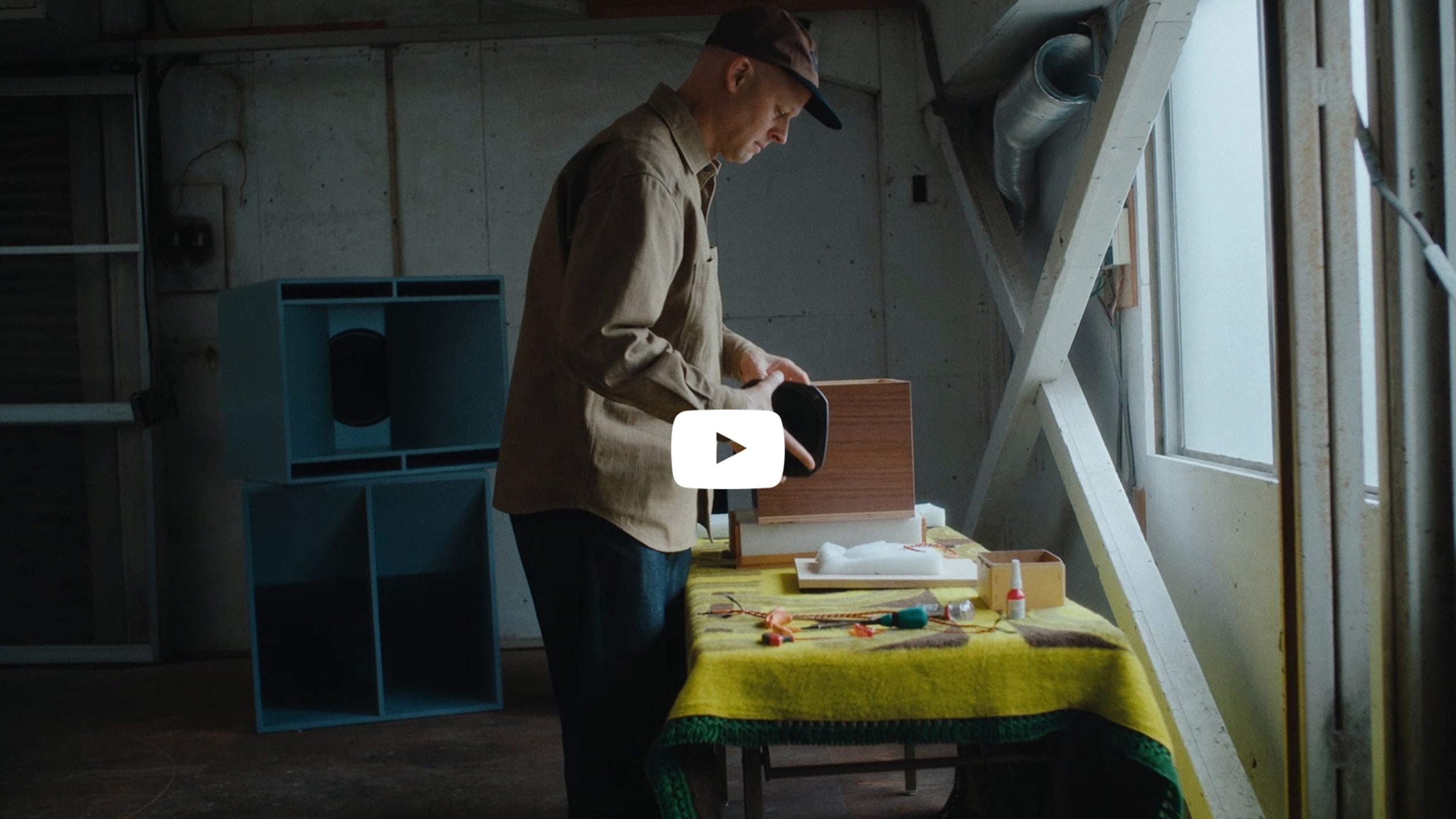 A man wearing a brown shirt and cap stands at a worktable arranging materials near a window in a studio space.