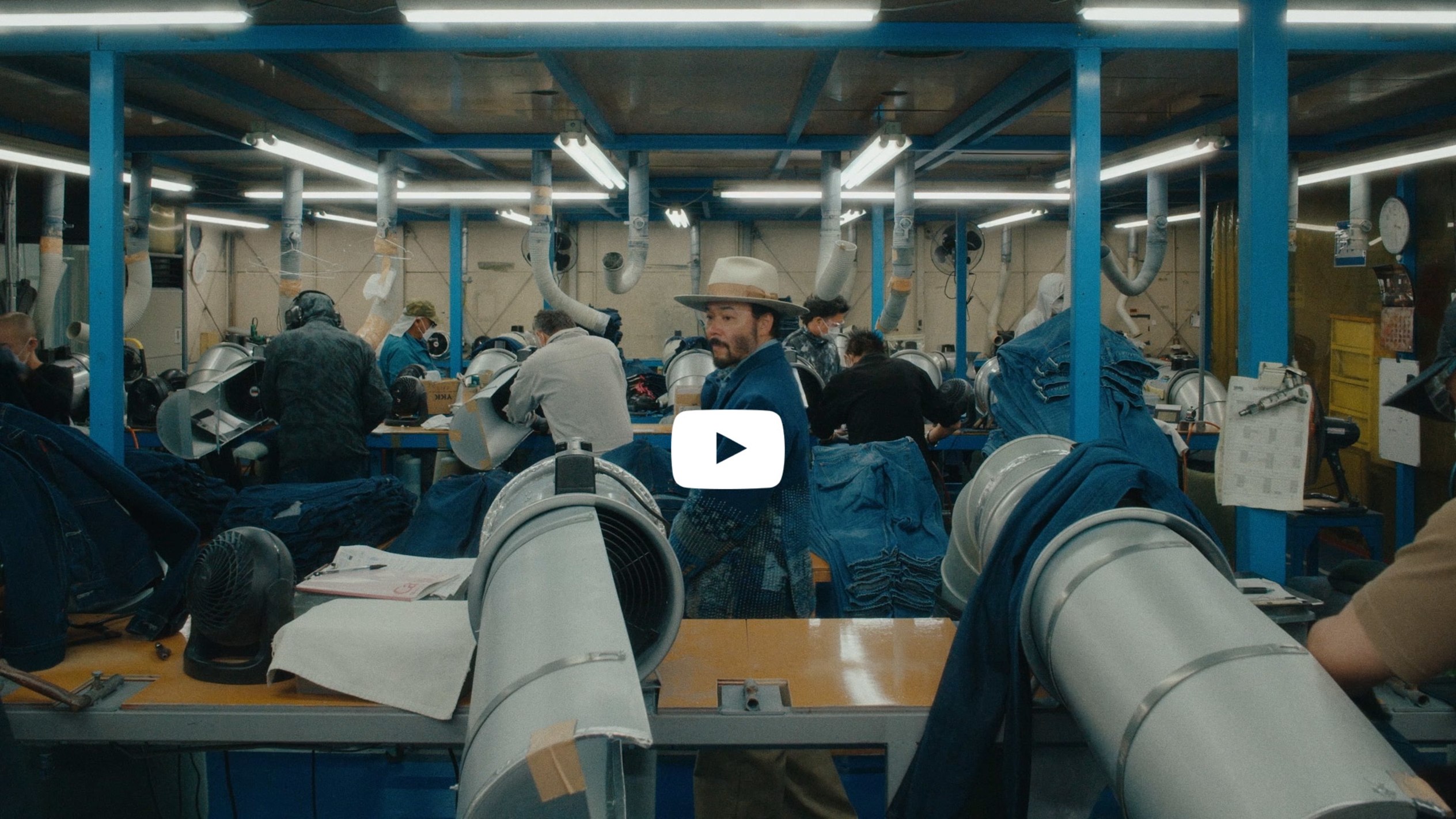 A man wearing a wide-brim hat and tailored jacket stands inside an industrial space, looking upward.