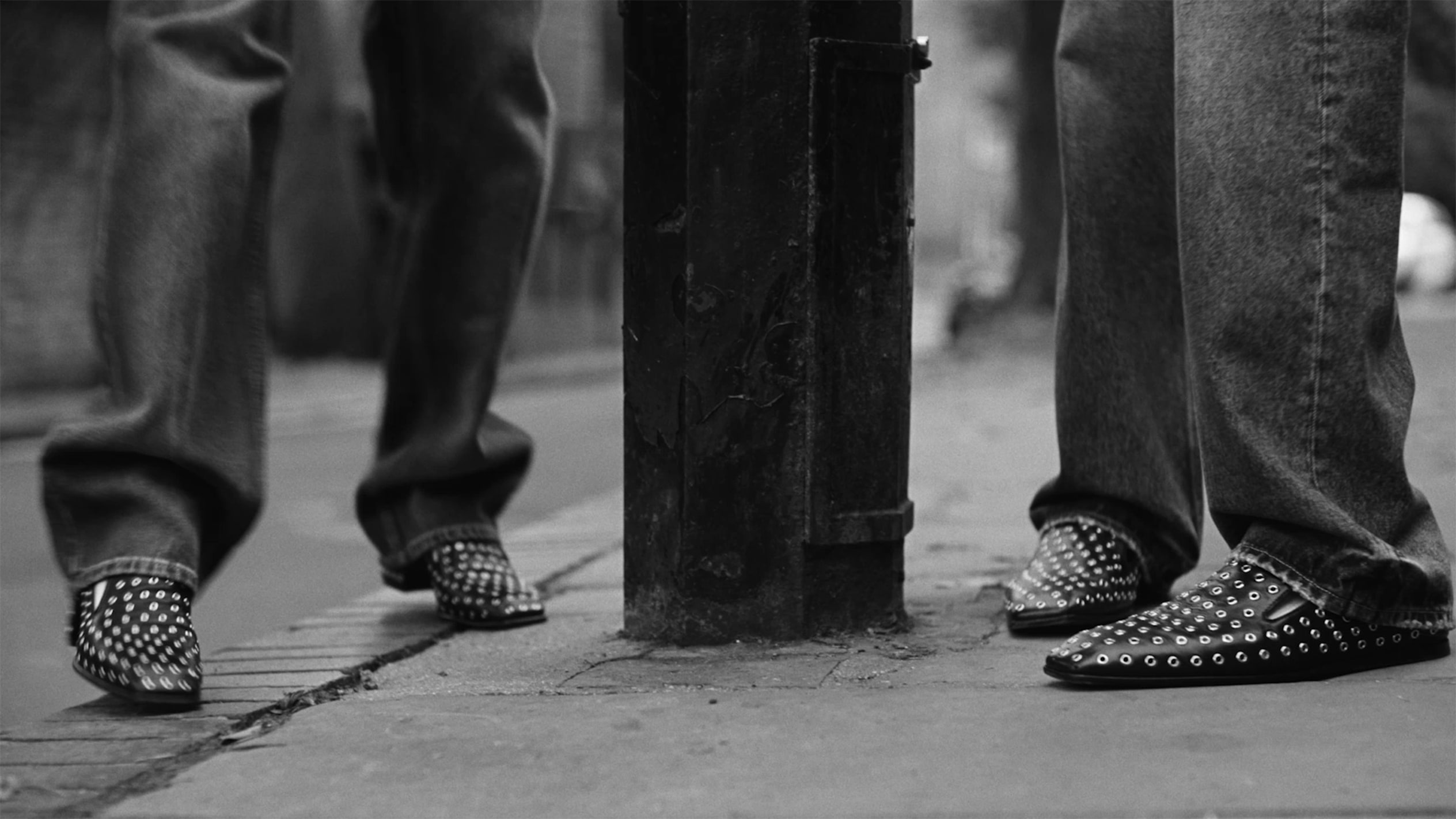 Black and white close-up image showing two people wearing studded black Eliot slippers with wide-leg jeans, photographed on a city pavement beside a worn lamppost.