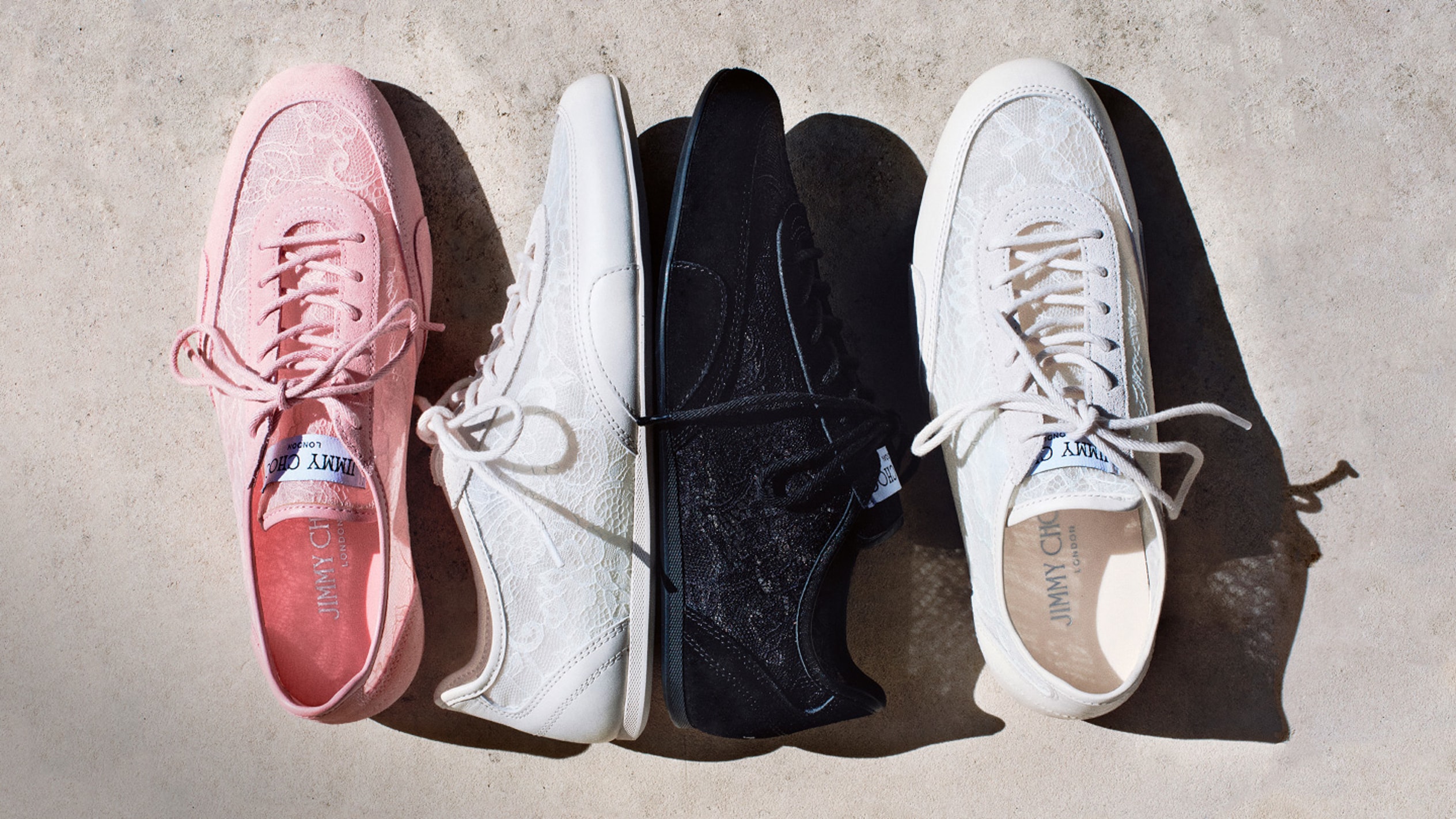 Four lace-detailed trainers laid side-by-side in rose, latte and black on a concrete surface.
