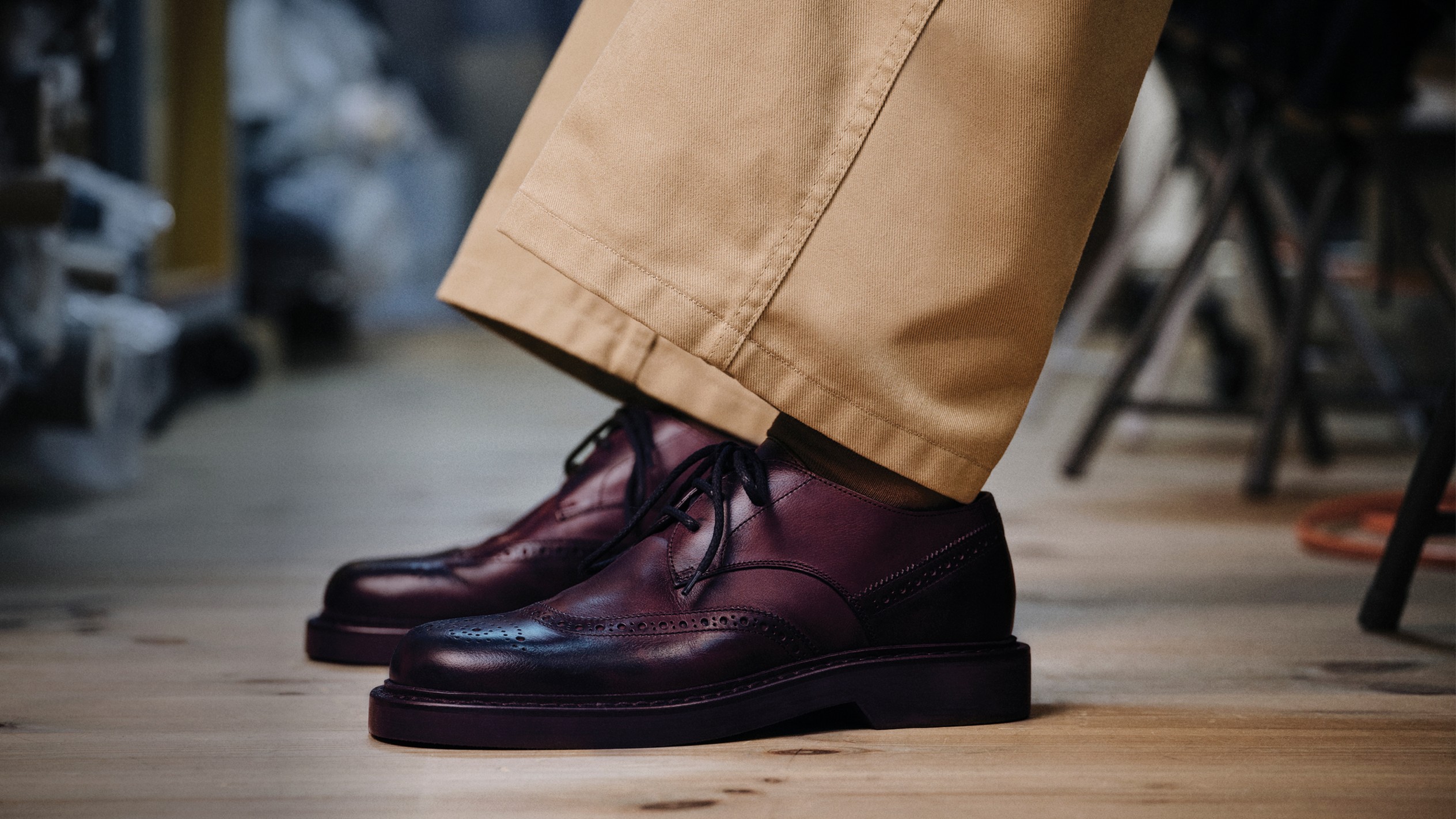 Brown leather Jimmy Choo men&rsquo;s lace-up shoes worn with beige trousers, photographed from the ankle down on a wooden floor.